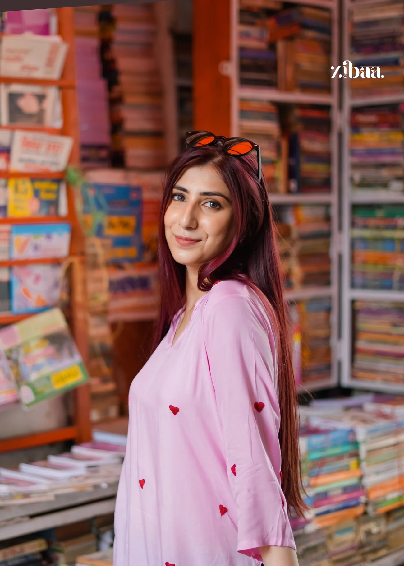 Wearing a pink chikankari kurti with heart motifs, the model poses on a street with a book stall visible in the background.