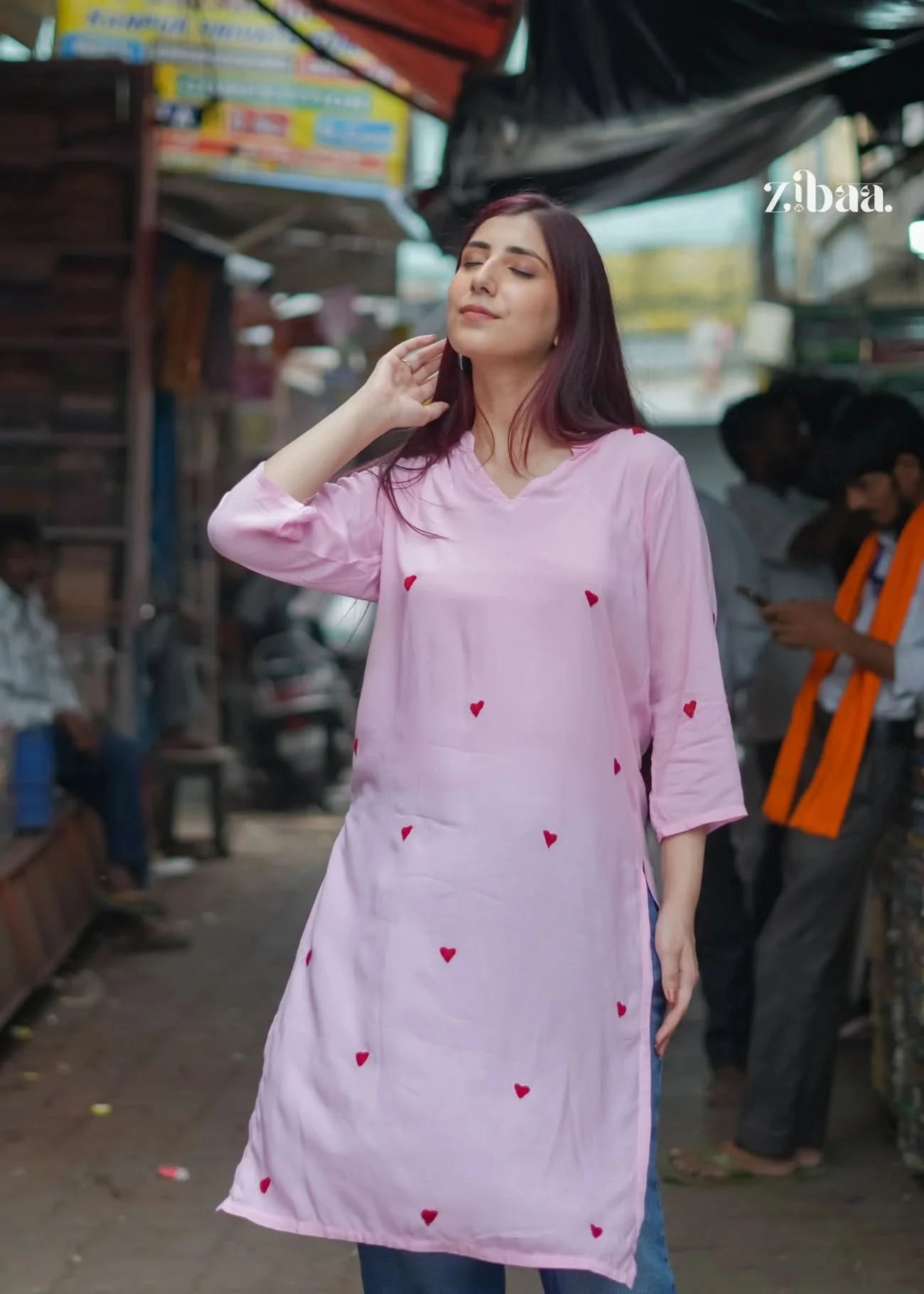 The model showcases a pink chikankari kurti with red heart embroidery in a casual front pose on a bustling street.