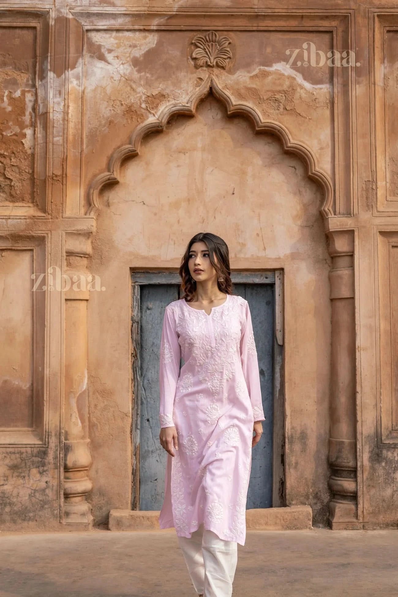 Model dressed in a pastel pink Lucknowi Kurti with white embroidery, posing in front of an ornate arched doorway in a heritage setting