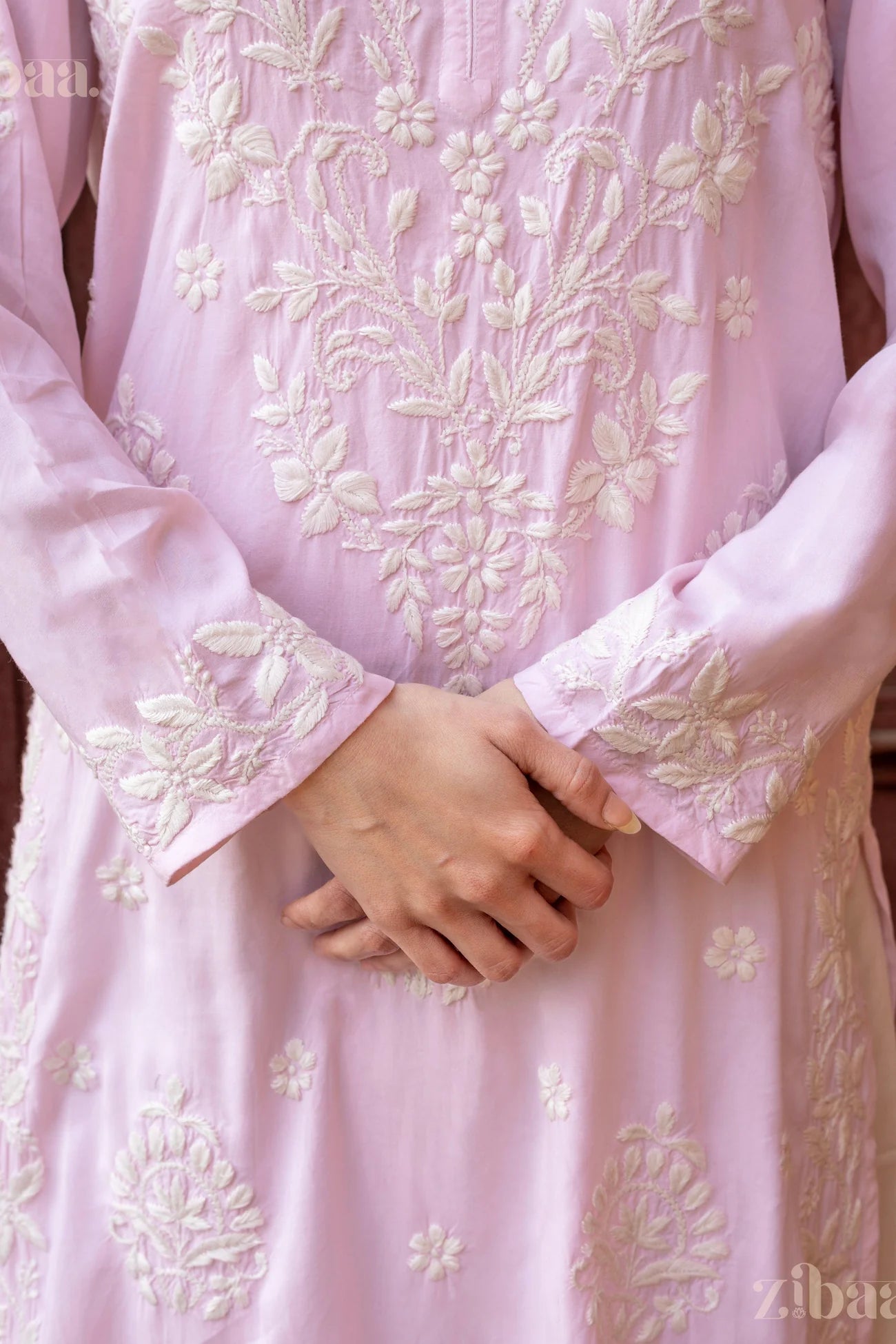 Close-up of hands folded gracefully over a pastel pink Chikankari Kurti, showcasing intricate floral embroidery on the sleeves and bodice