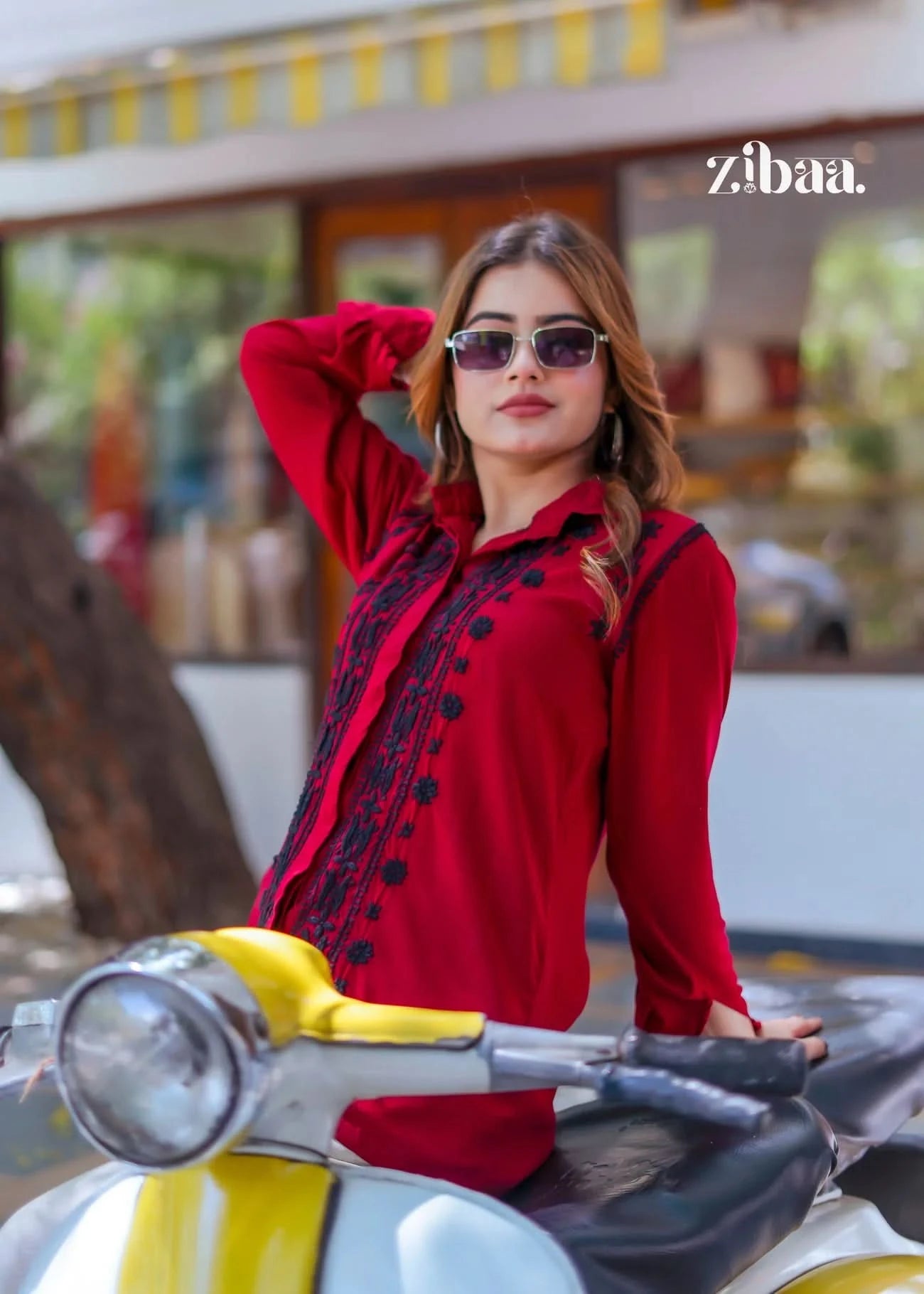 The model strikes a pose on a scooter, wearing a red chikankari shirt adorned with fine black embroidery.