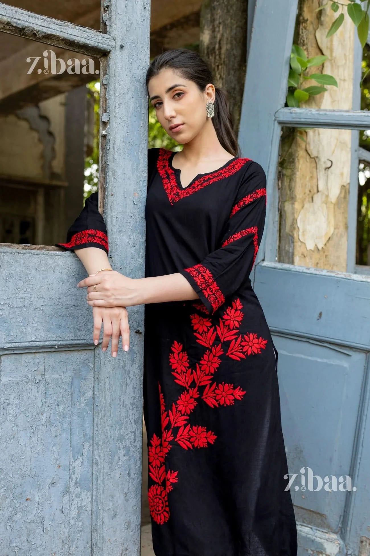 Woman wearing a black dress with red embroidery leaning against a wall.