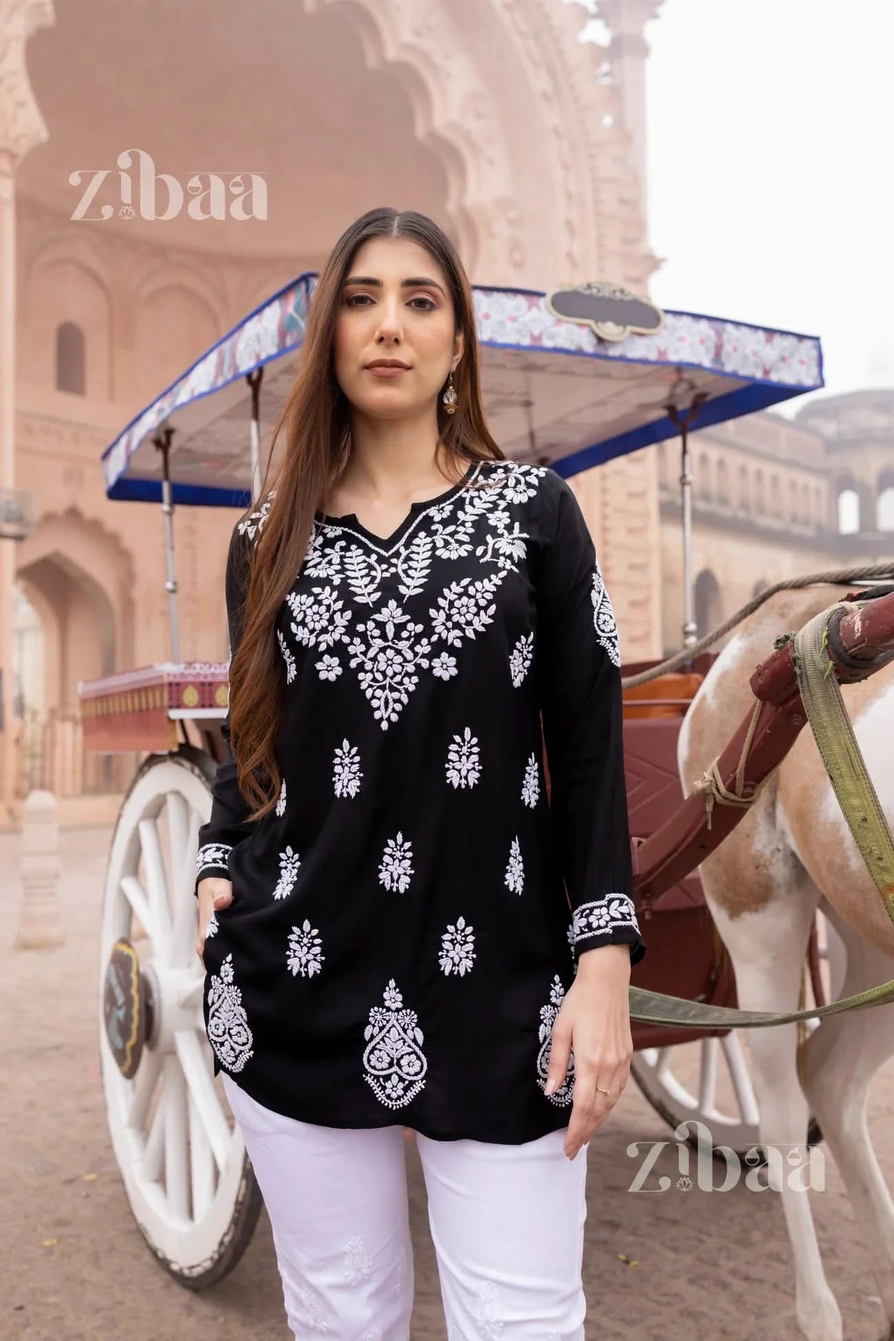 A woman wearing a black short chikankari kurti with floral embroidery, standing in front of a traditional rickshaw.