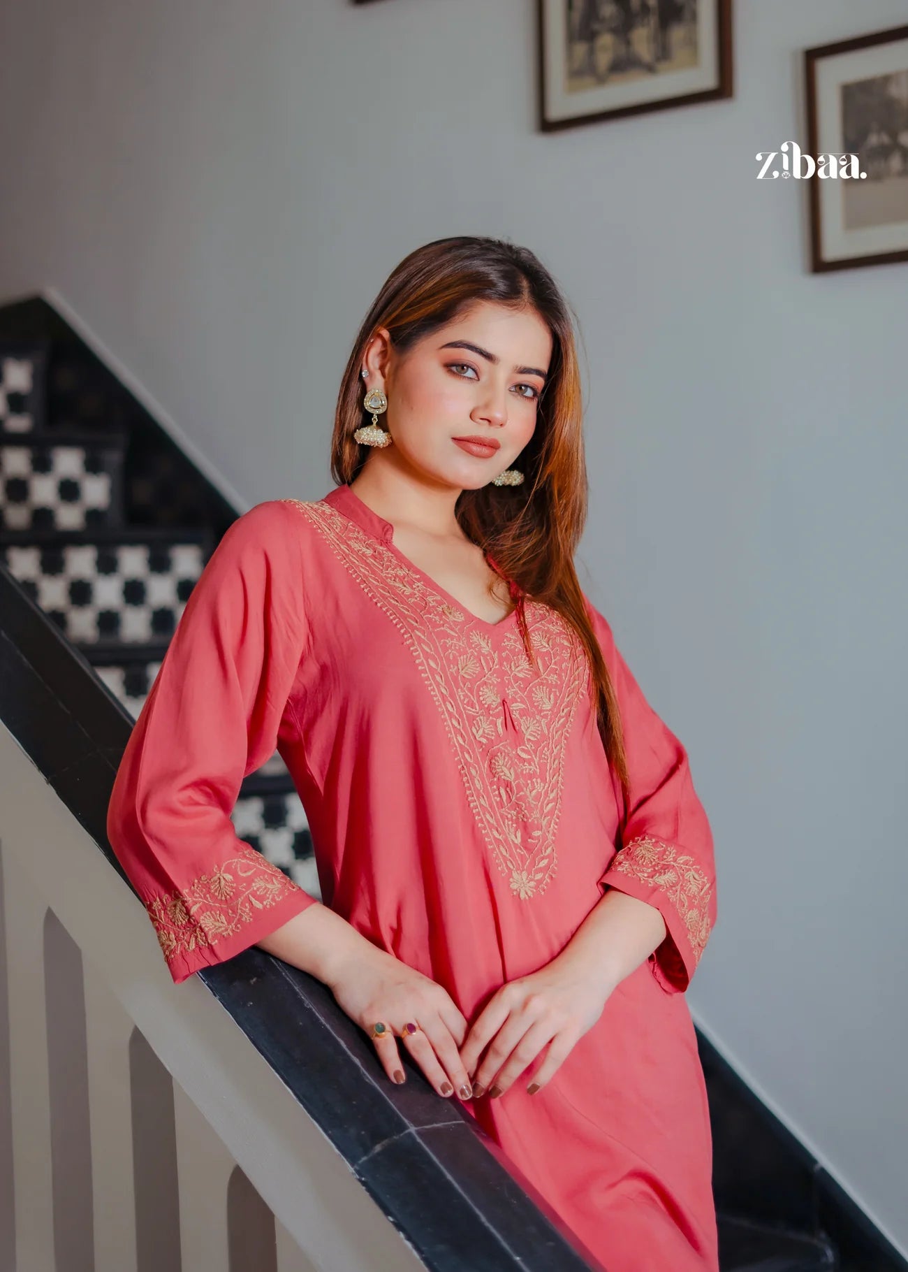 The model poses on a staircase wearing a coral chikankari kurti, with the intricate golden embroidery adding a touch of elegance.