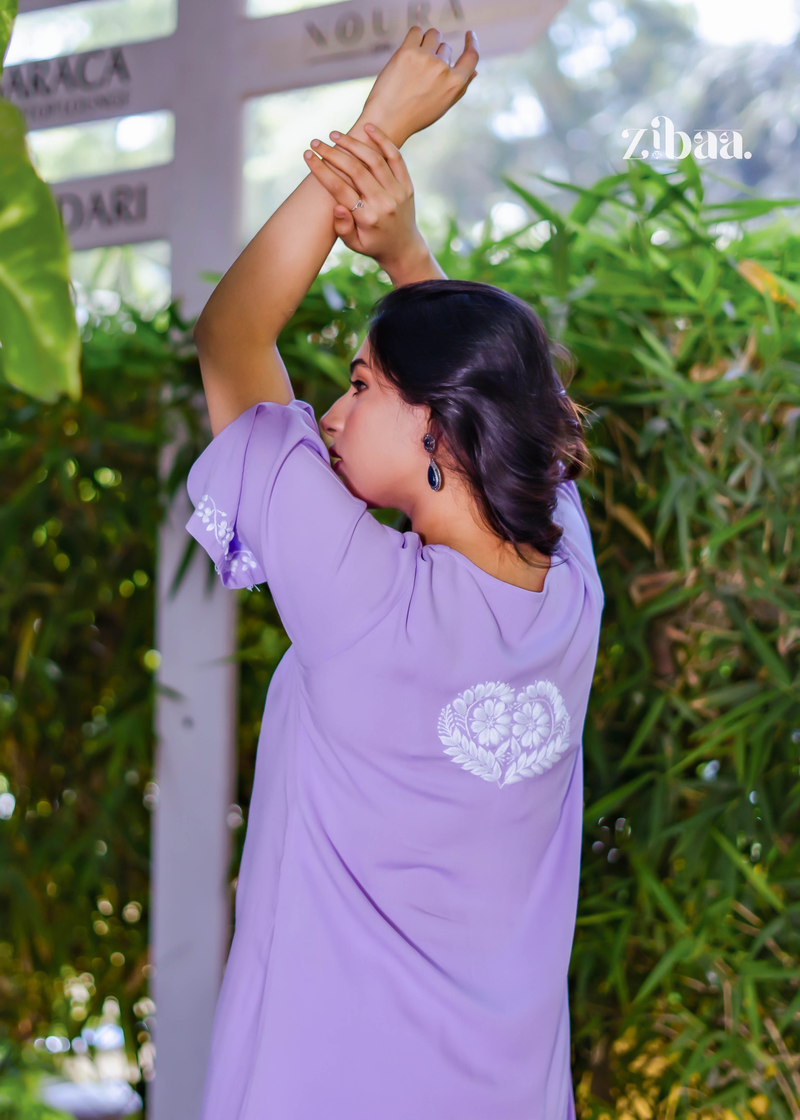 The model raises her hands, showcasing the back embroidery of the lavender chikankari kurti, with the soft lavender tone standing out in the outdoor setting.