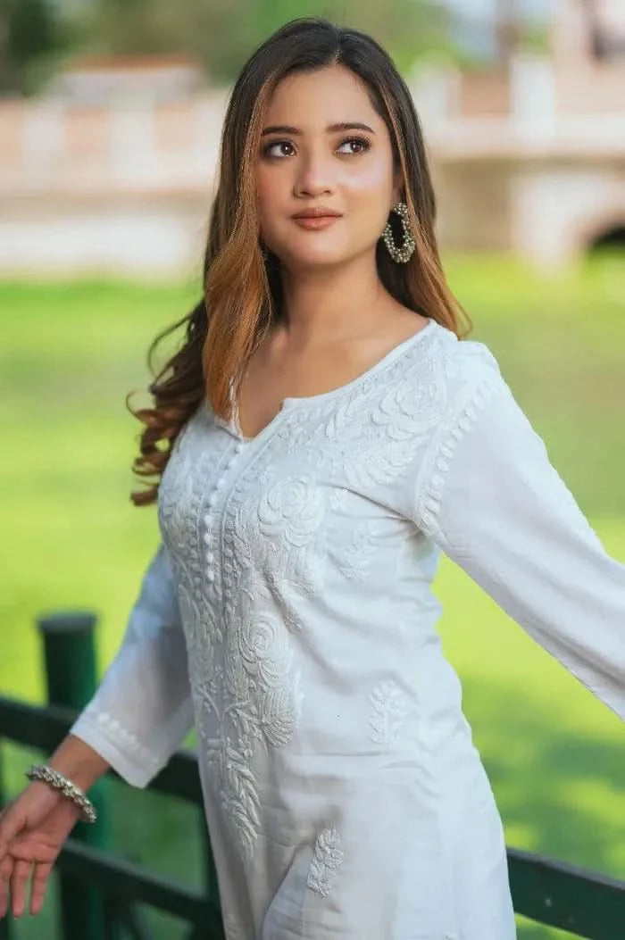 A woman wearing a white Chikankari kurti with floral embroidery stands near a railing looking upward with a soft expression.