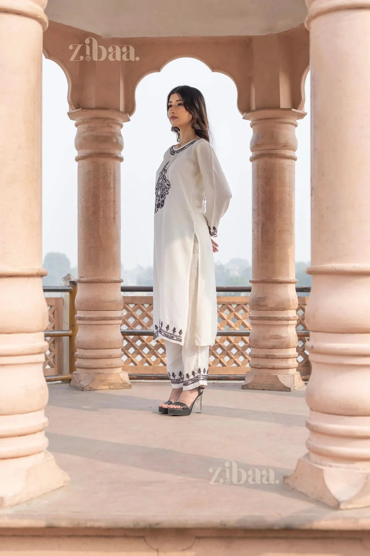 A woman wearing a white Chikankari kurti with black embroidery stands between ornate pillars, gazing into the distance outdoors.