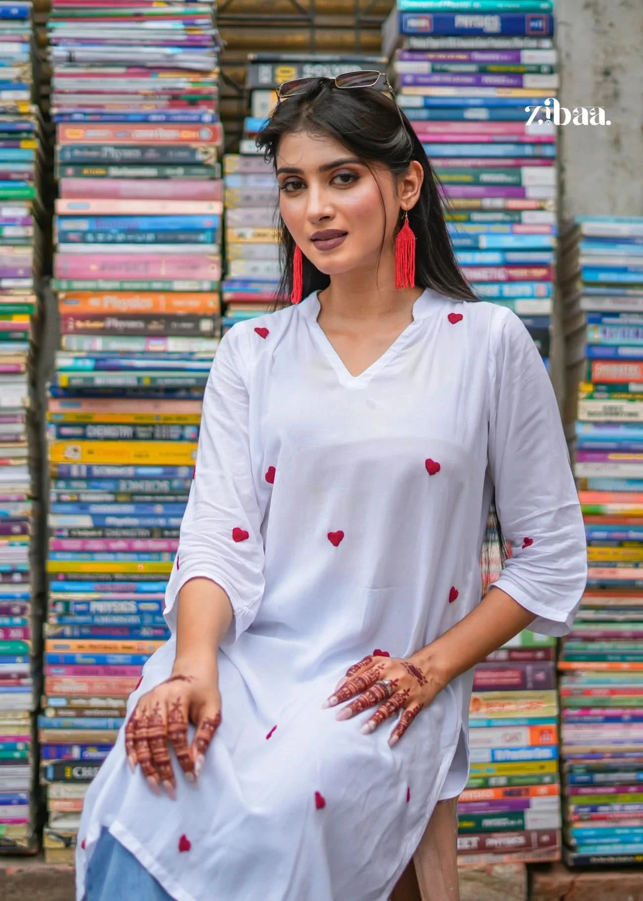 The model sits comfortably on a chair, wearing a white chikankari kurti, with a book stall softly visible in the background, blending naturally into the street scene.