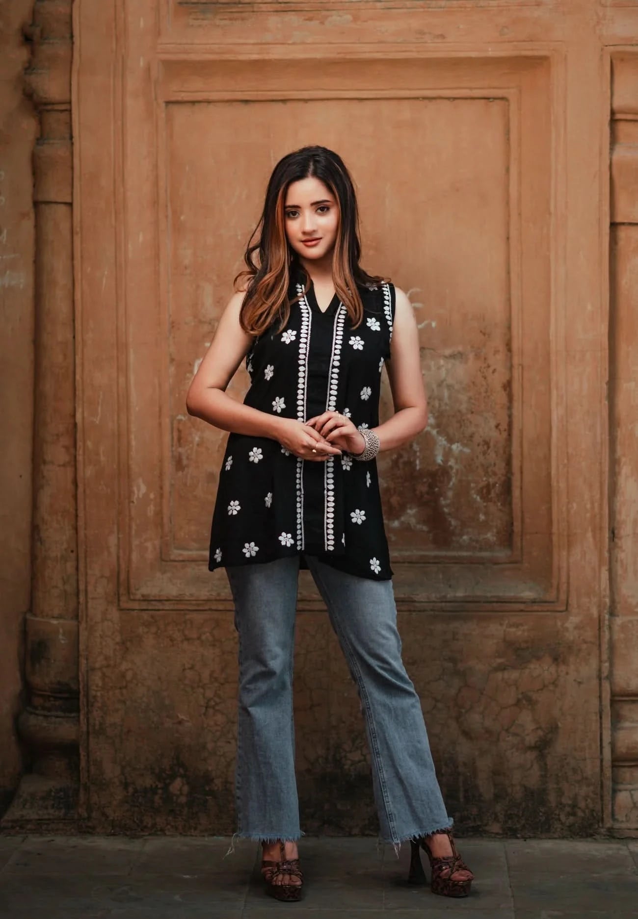 A woman wearing a sleeveless black Chikankari shorty kurti with white floral embroidery stands with, arms crossed, against a rustic wall.