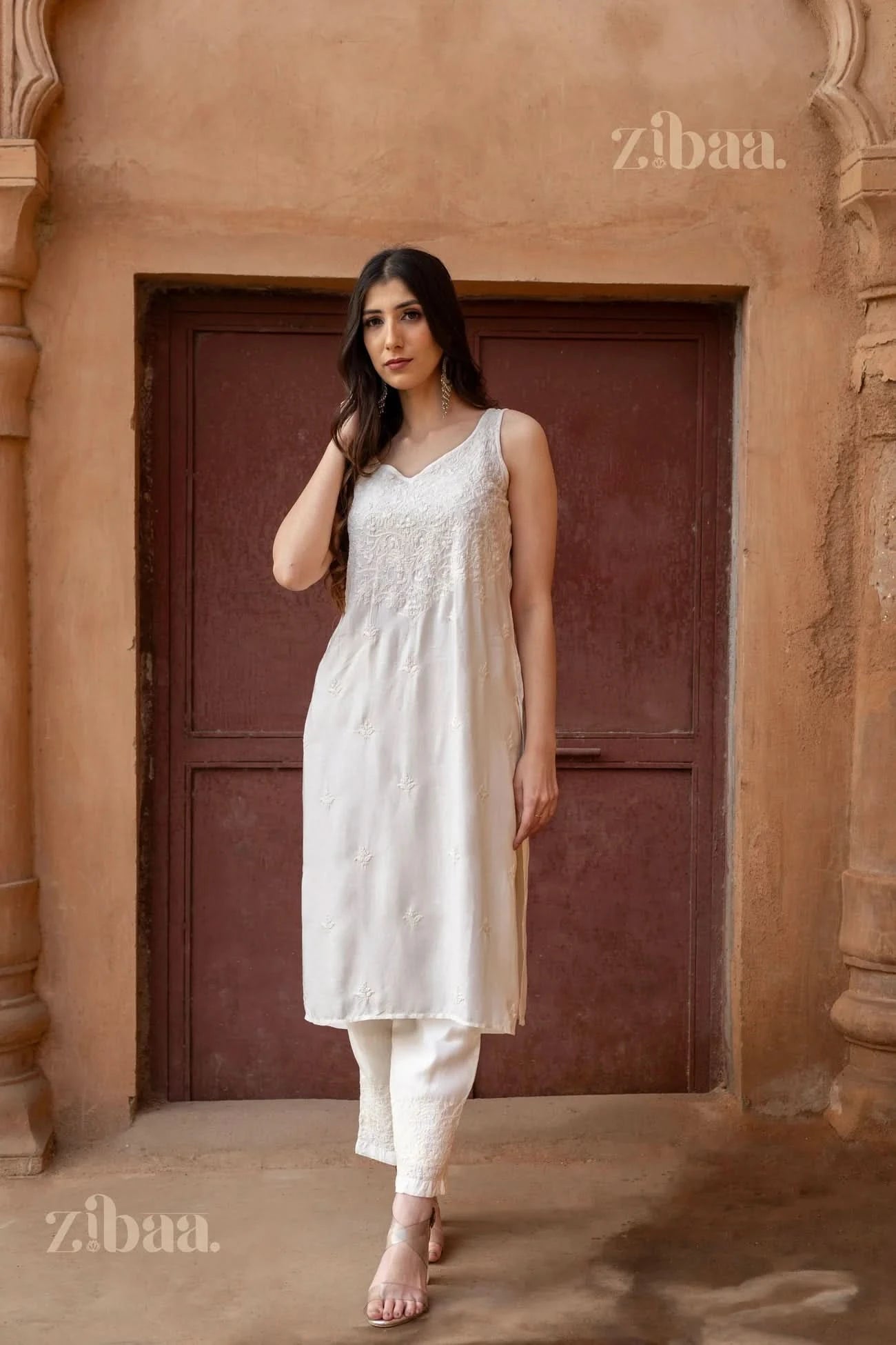 Model wearing a sleeveless Chikankari Kurta Set in white, featuring delicate embroidery, posing in a rustic architectural setting.