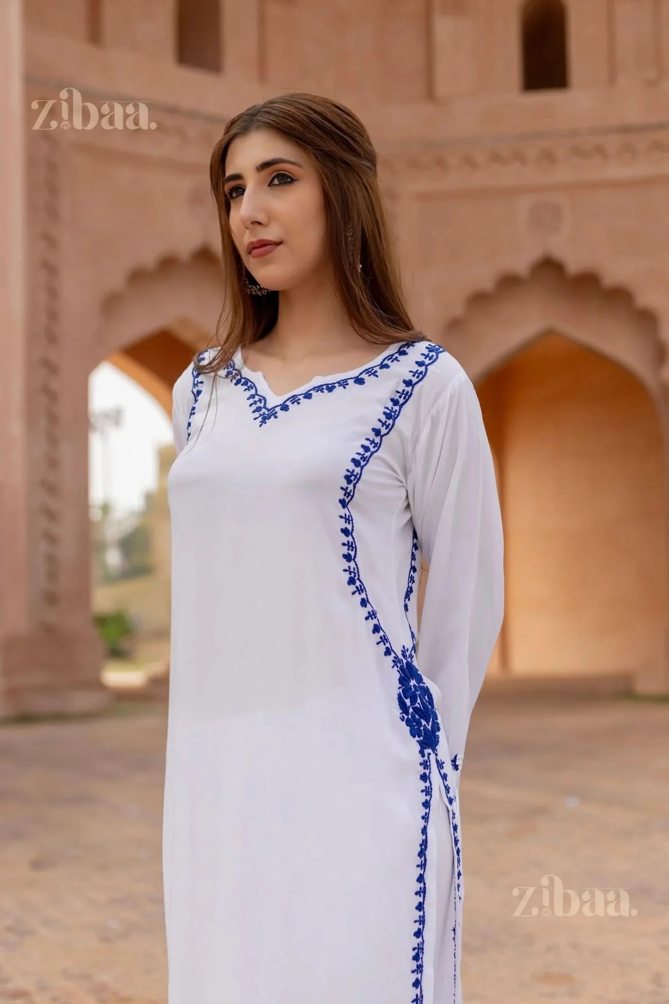 Woman wearing a white dress with blue embroidery in front of an architectural background