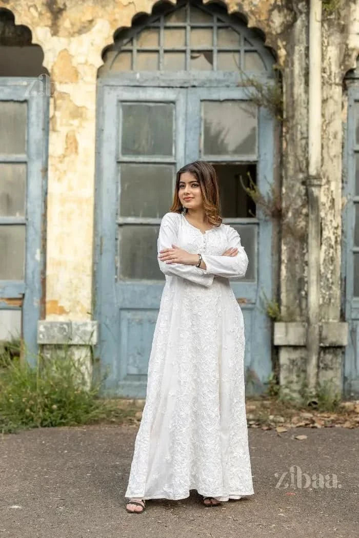 A woman wearing a white Chikankari Anarkali with intricate floral embroidery stands with arms crossed, posing by a rustic blue door.