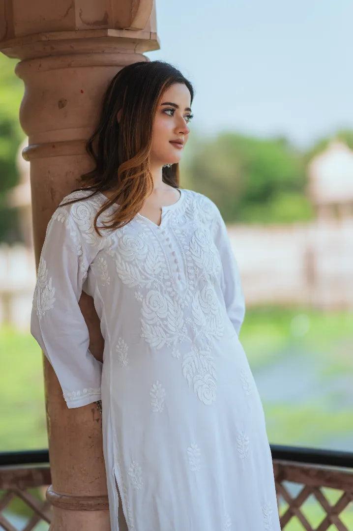 A woman wearing a white Chikankari kurti with intricate embroidery leans softly against a pillar, gazing thoughtfully into the distance.