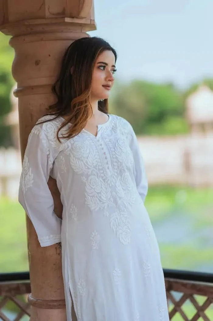 A woman wearing a white Chikankari kurti with intricate embroidery leans softly against a pillar, gazing thoughtfully into the distance.