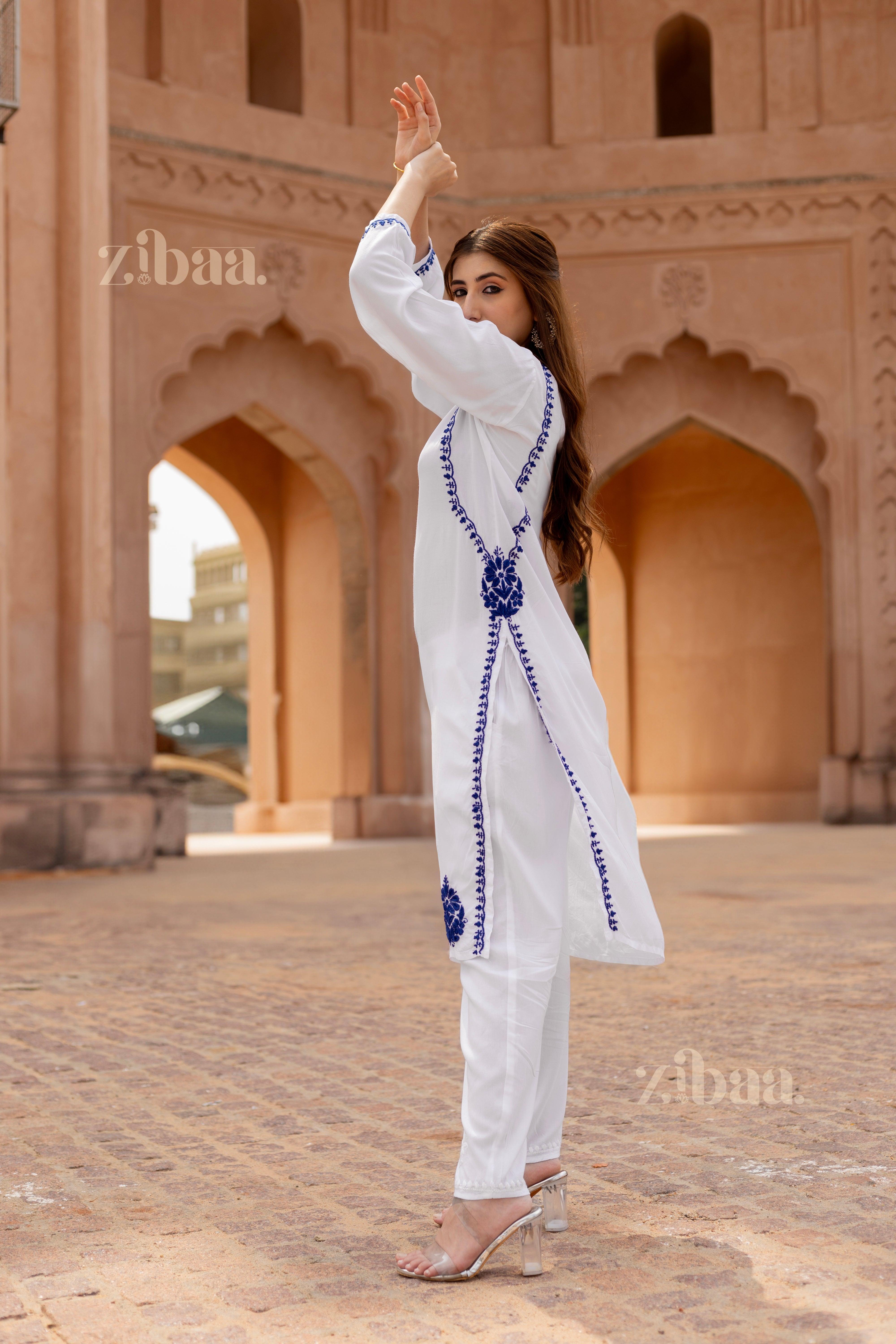 Woman gracefully posing with arms raised, wearing a white kurti for women adorned with blue embroidery, standing in a heritage setting