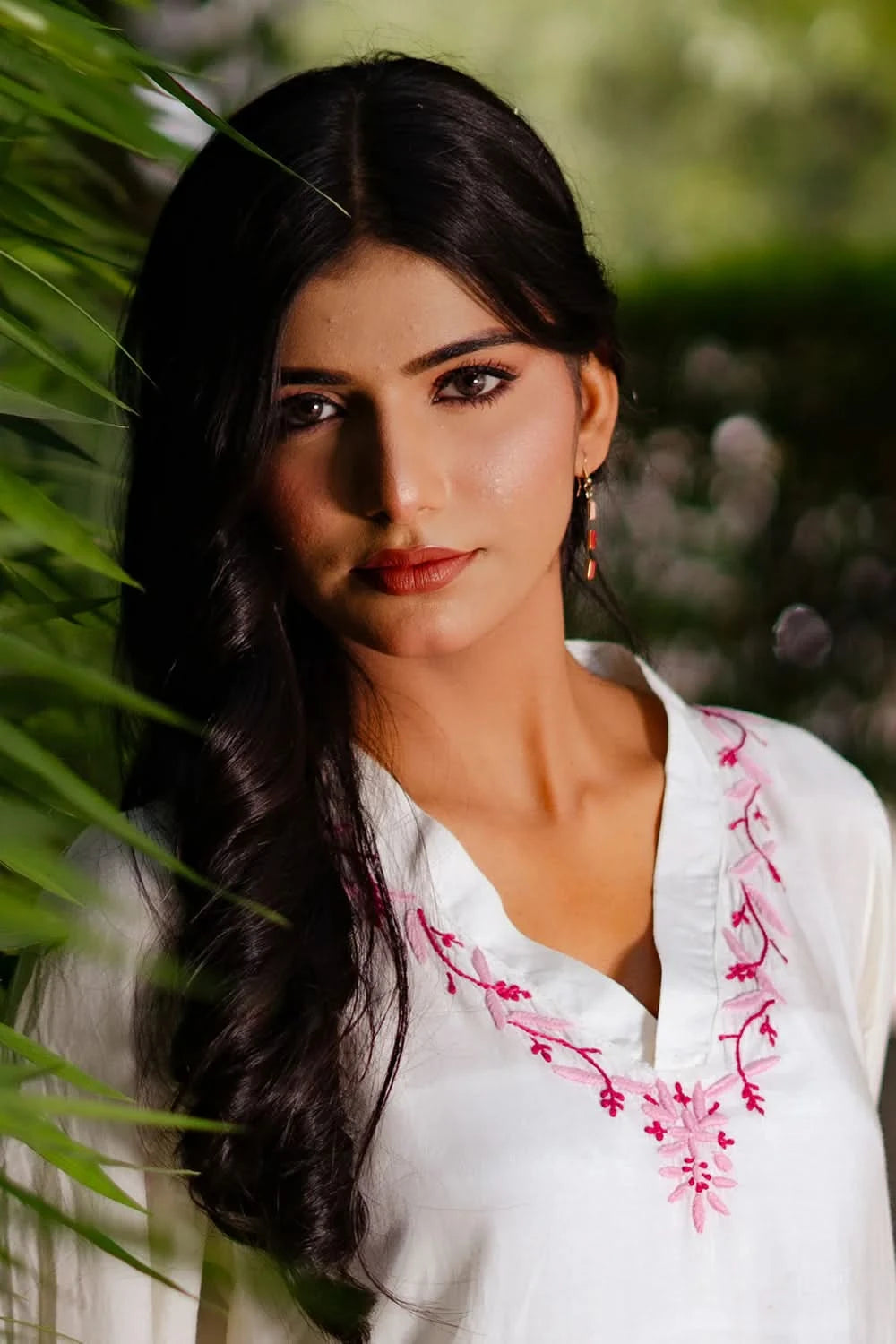Woman wearing a white chikankari kurti with pink embroidery, standing among green foliage.