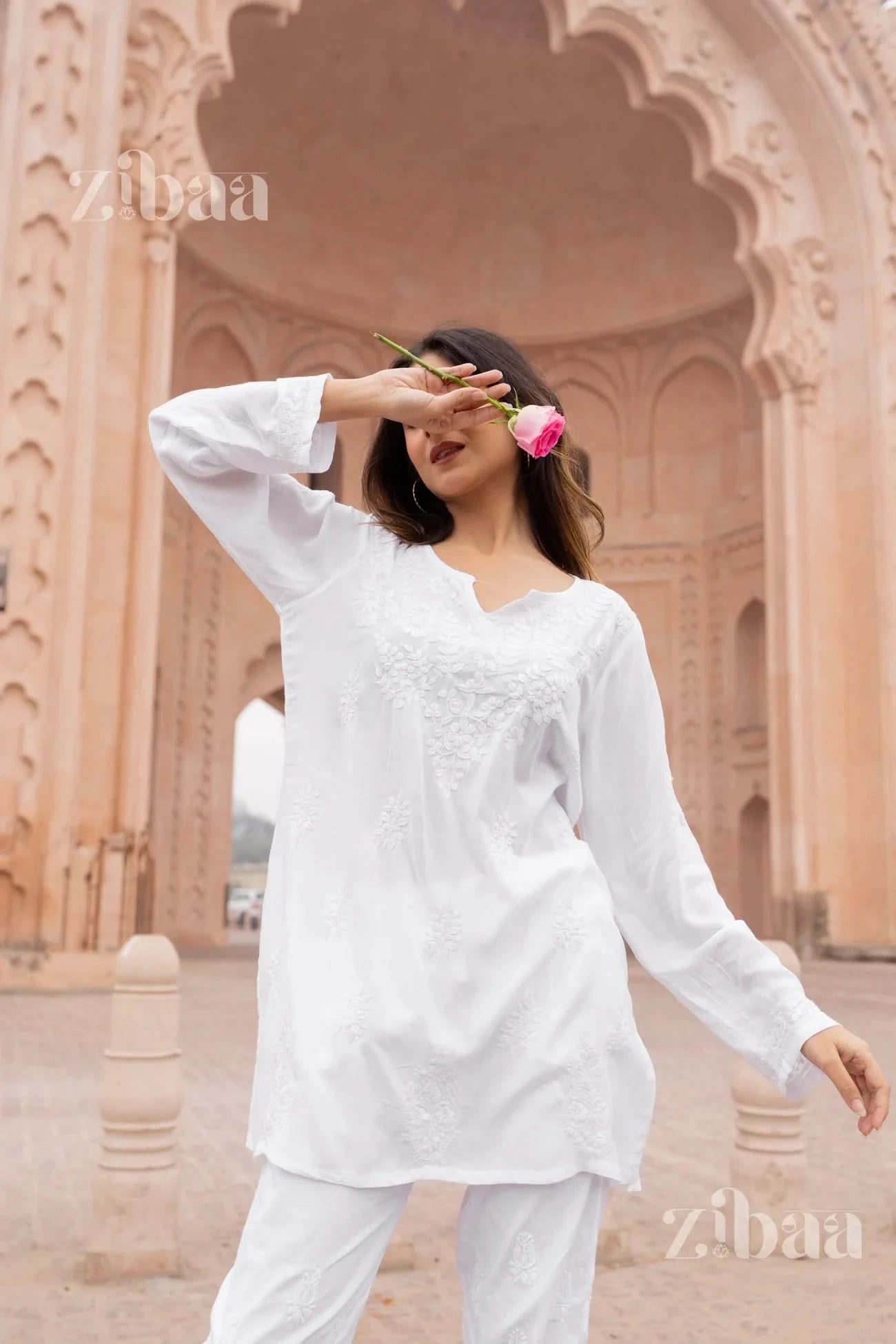Woman in a white outfit holding a pink flower in front of an architectural background with 'Zibaa' branding.