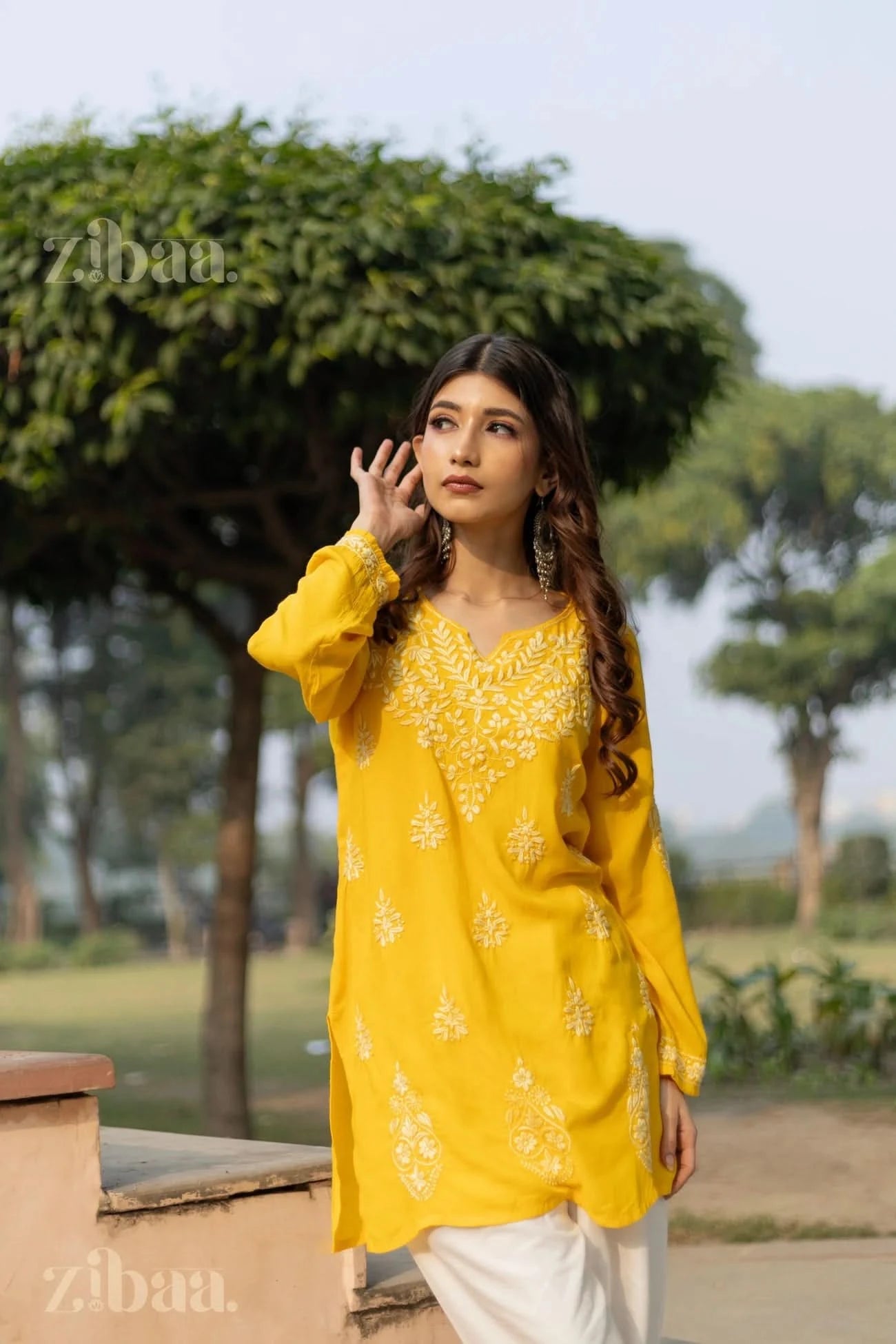 A woman wearing a yellow short kurti with white floral embroidery stands outdoors, touching her hair with one hand, gazing thoughtfully.