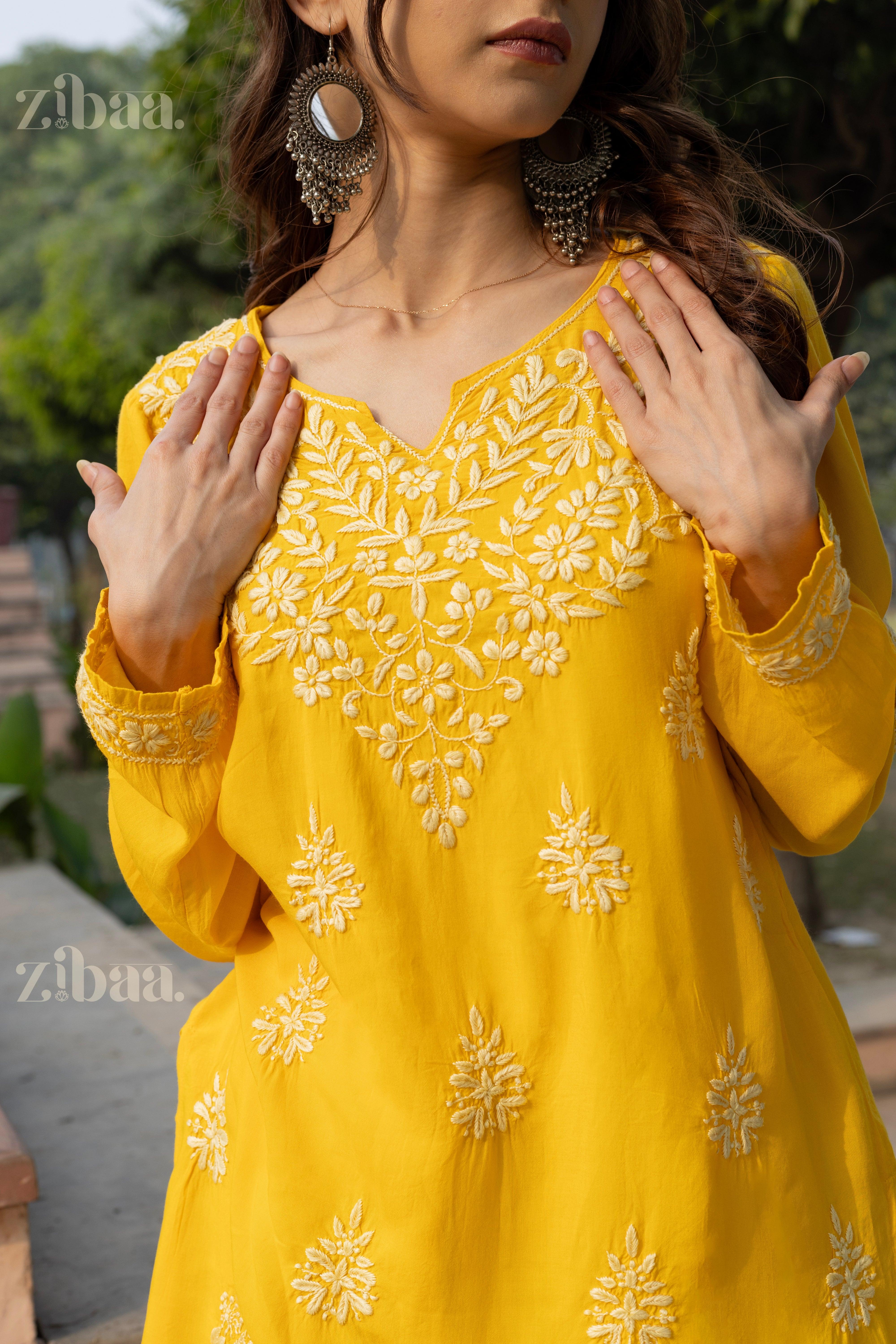 A woman wearing a yellow Chikankari kurti with intricate floral embroidery gently places her hands near the neckline, posing outdoors.