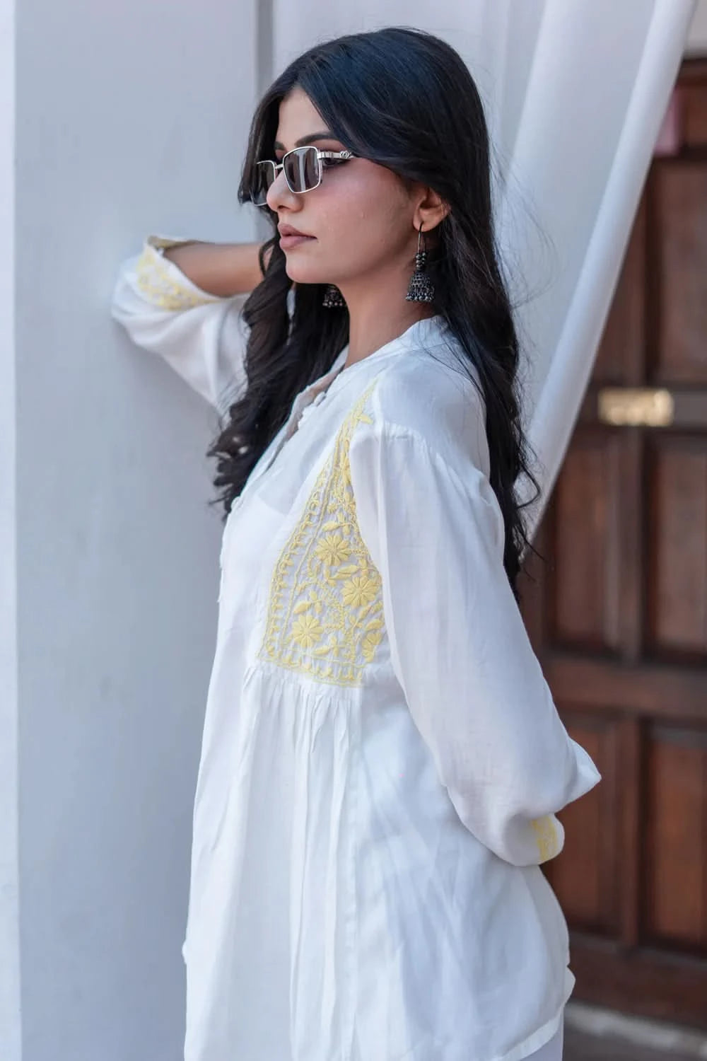 Woman wearing a white outfit with yellow embroidery, standing against a neutral background.