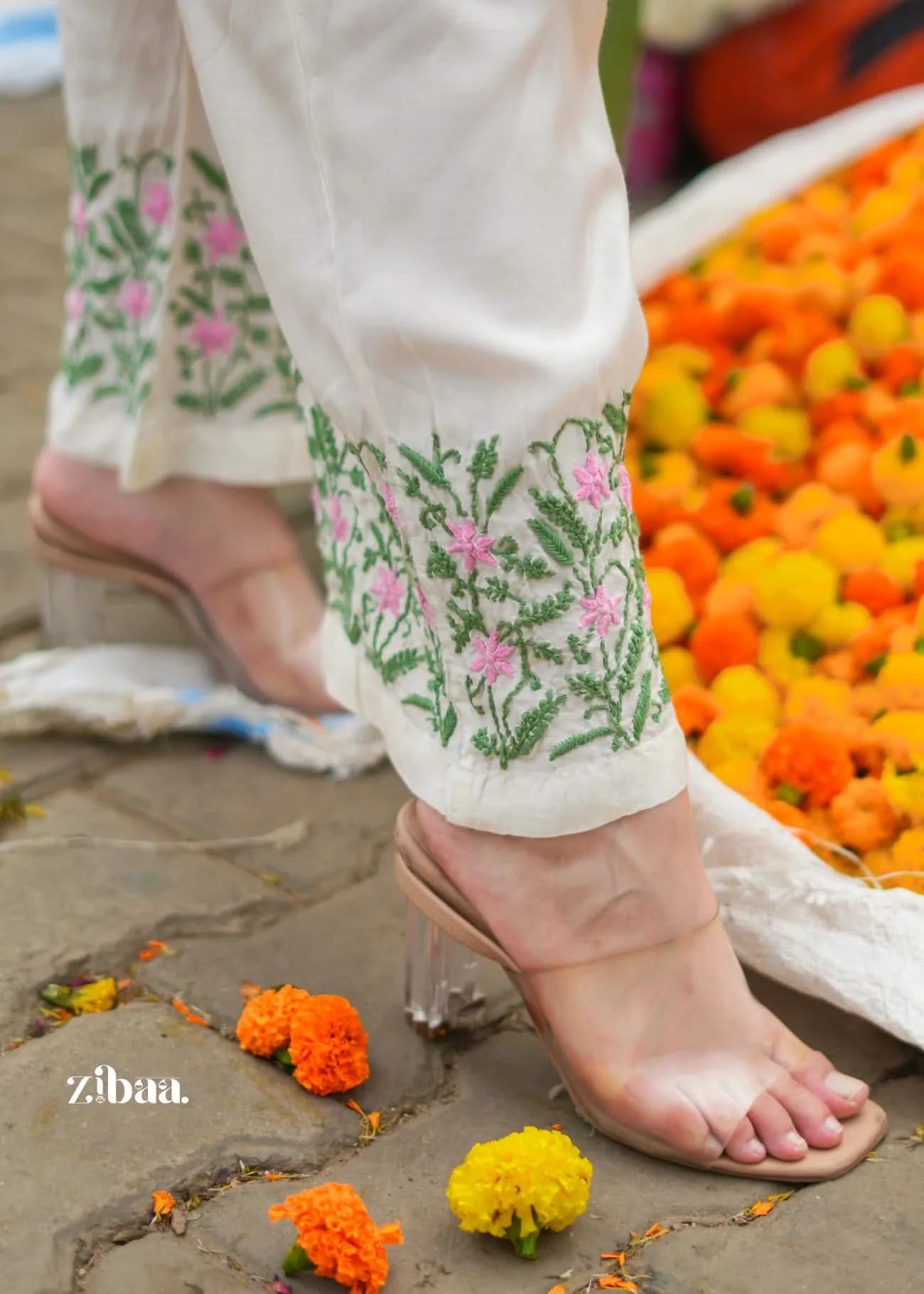 Person wearing white pants with floral embroidery and sandals, standing on a stone surface with flowers around.