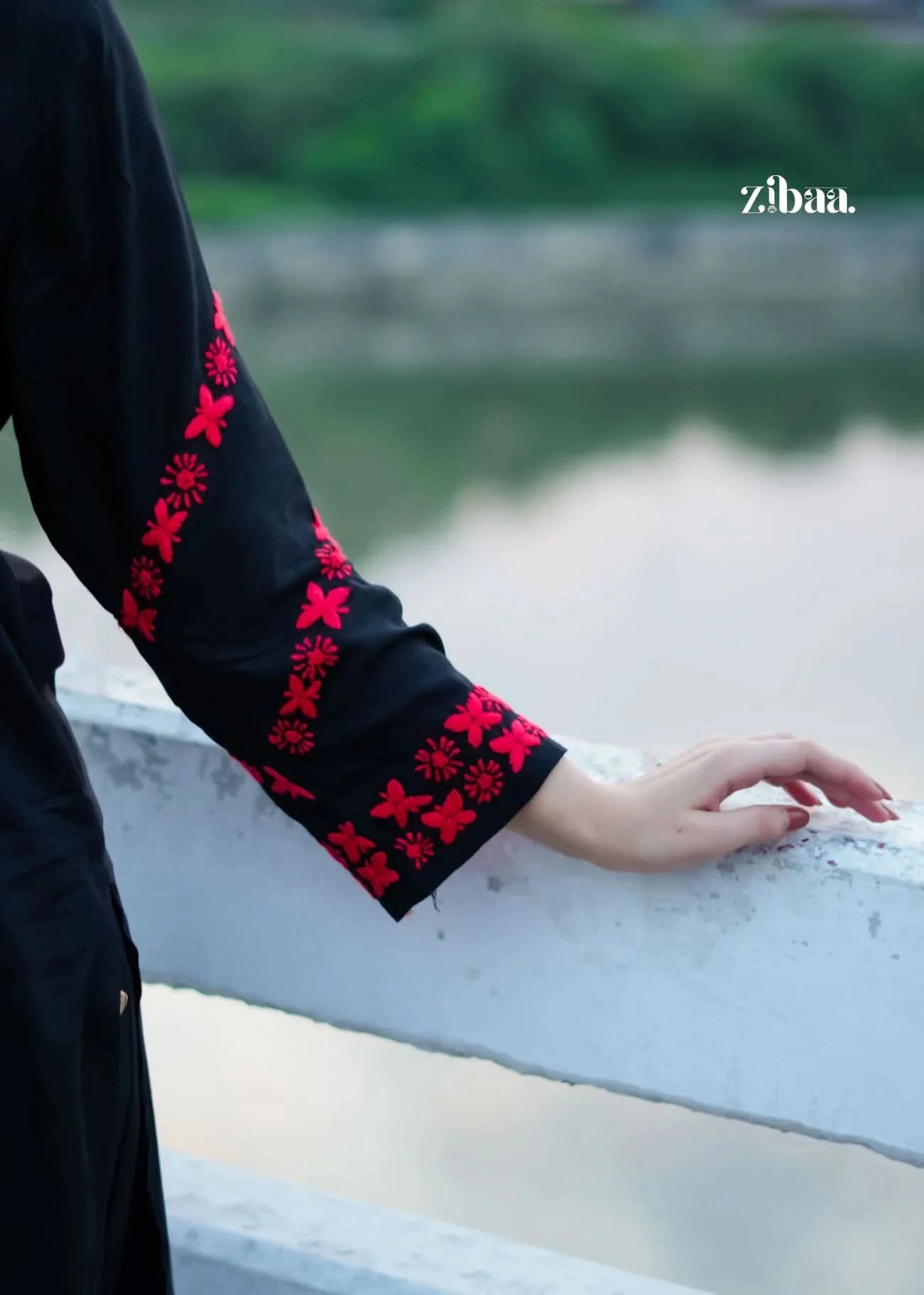 Person wearing a black outfit with red floral embroidery, standing by a body of water.