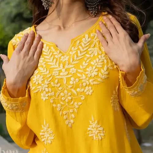 A girl wearing a yellow short kurti with intricate white Chikankari embroidery and earrings poses elegantly outdoors in natural light.