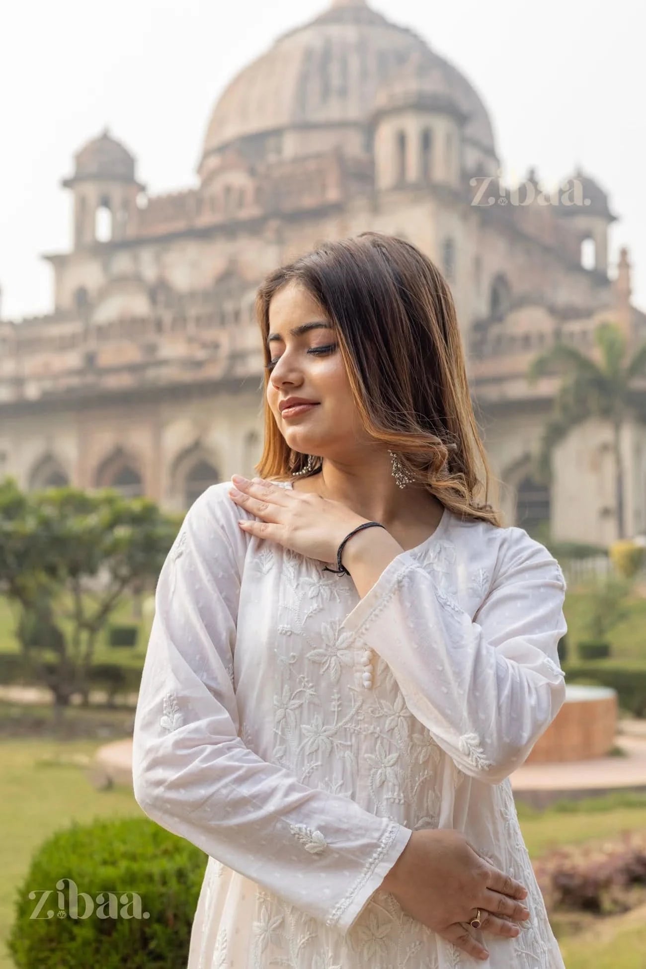 A woman in a white Chikankari Anarkali stands outdoors, softly touching her shoulder with a historic monument in the background.