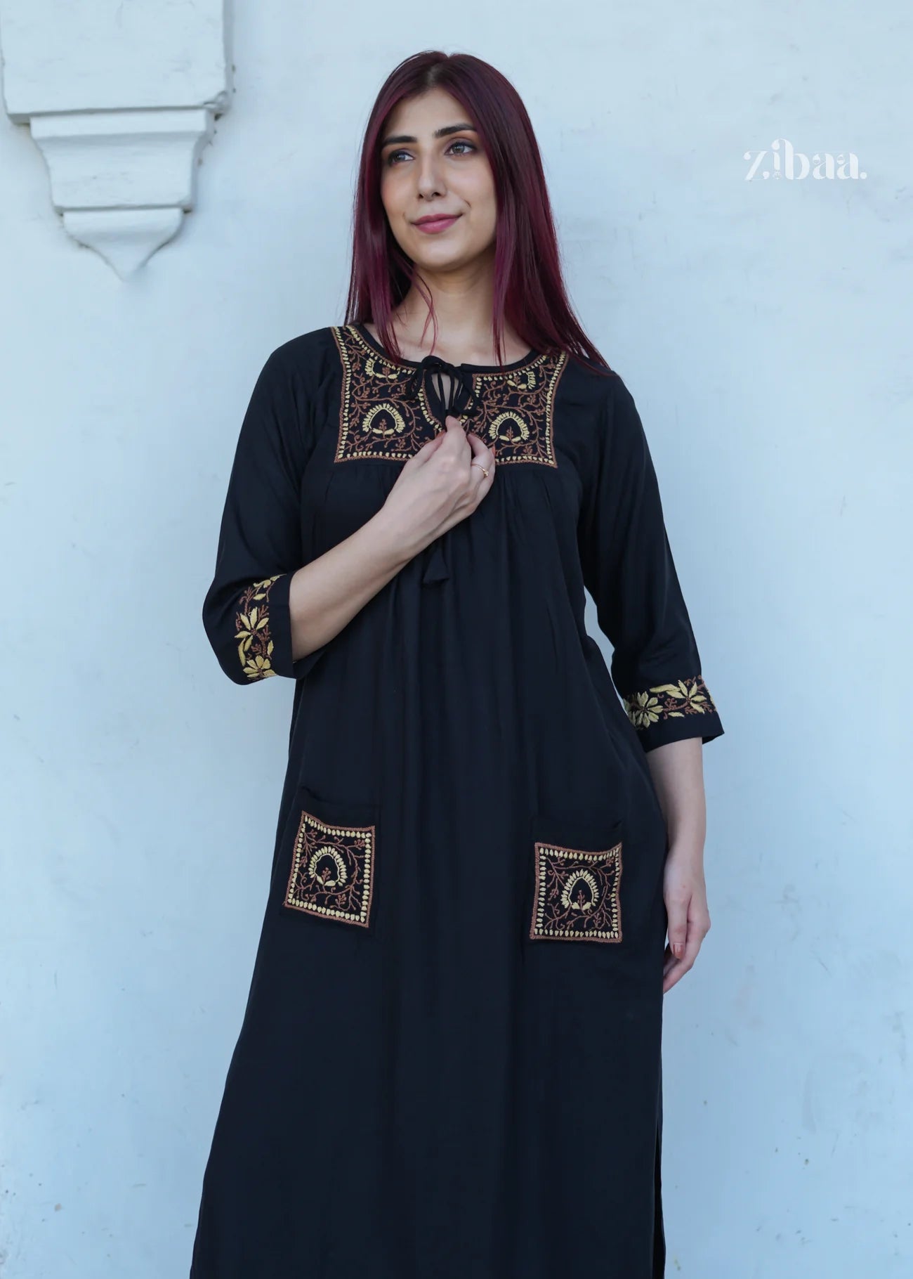 The model poses confidently in a black chikankari kurti with intricate golden threadwork, set against a clean white wall.