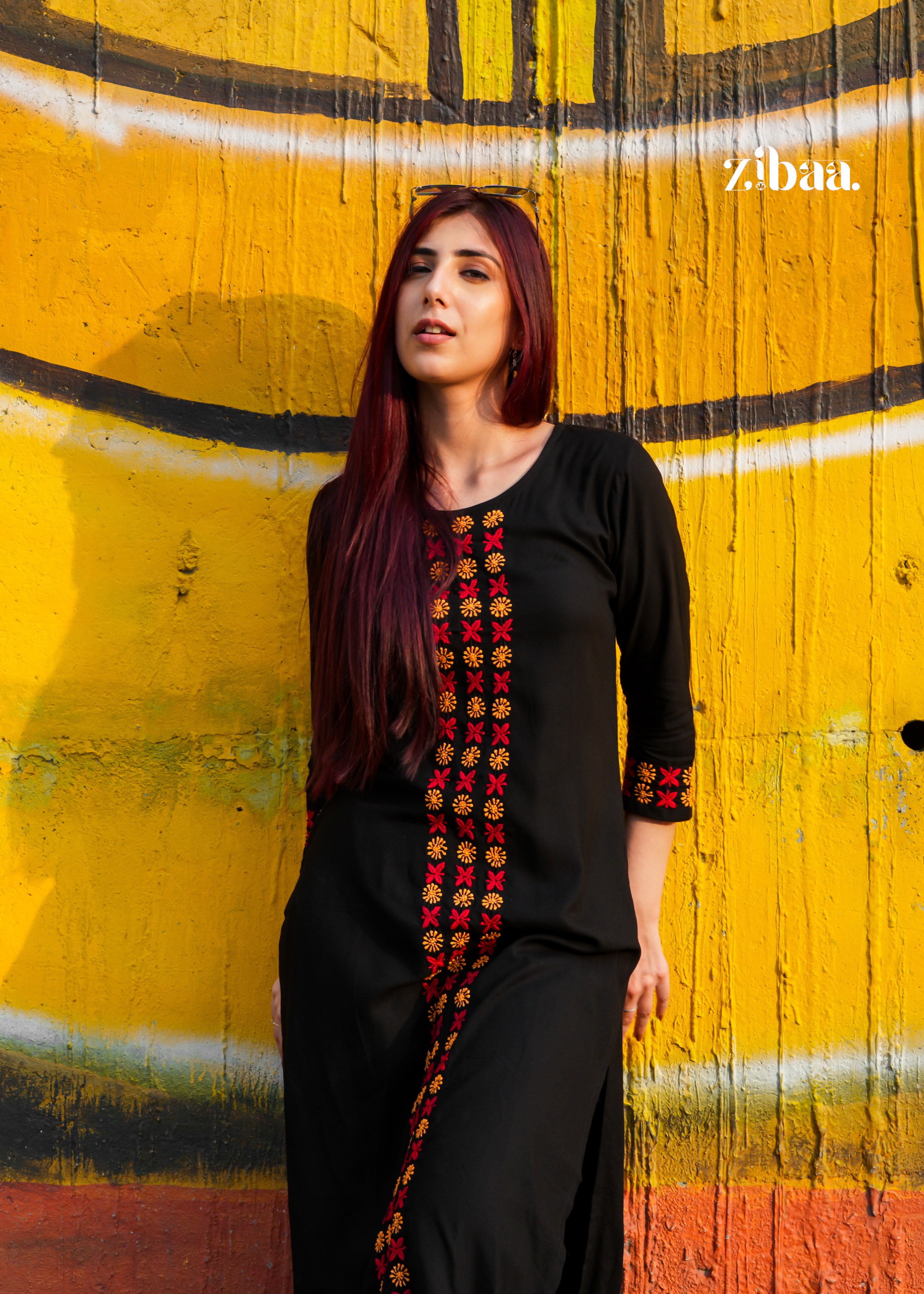 Woman in a black chikankari kurti set with colorful patterns standing against a yellow wall with graffiti.