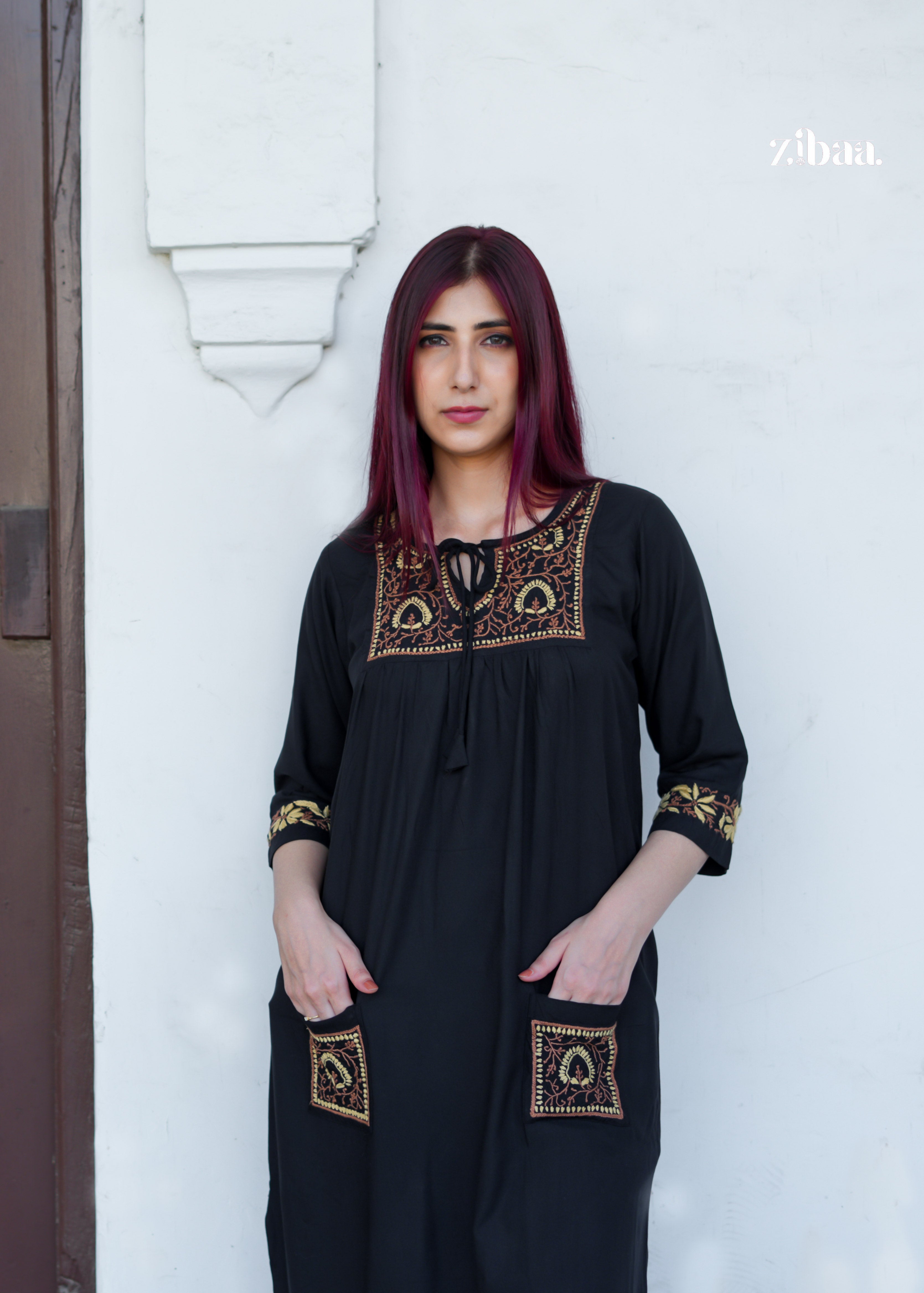 Standing in front of a white wall, the model shows off the intricate golden threadwork on her black chikankari kurti with effortless style.
