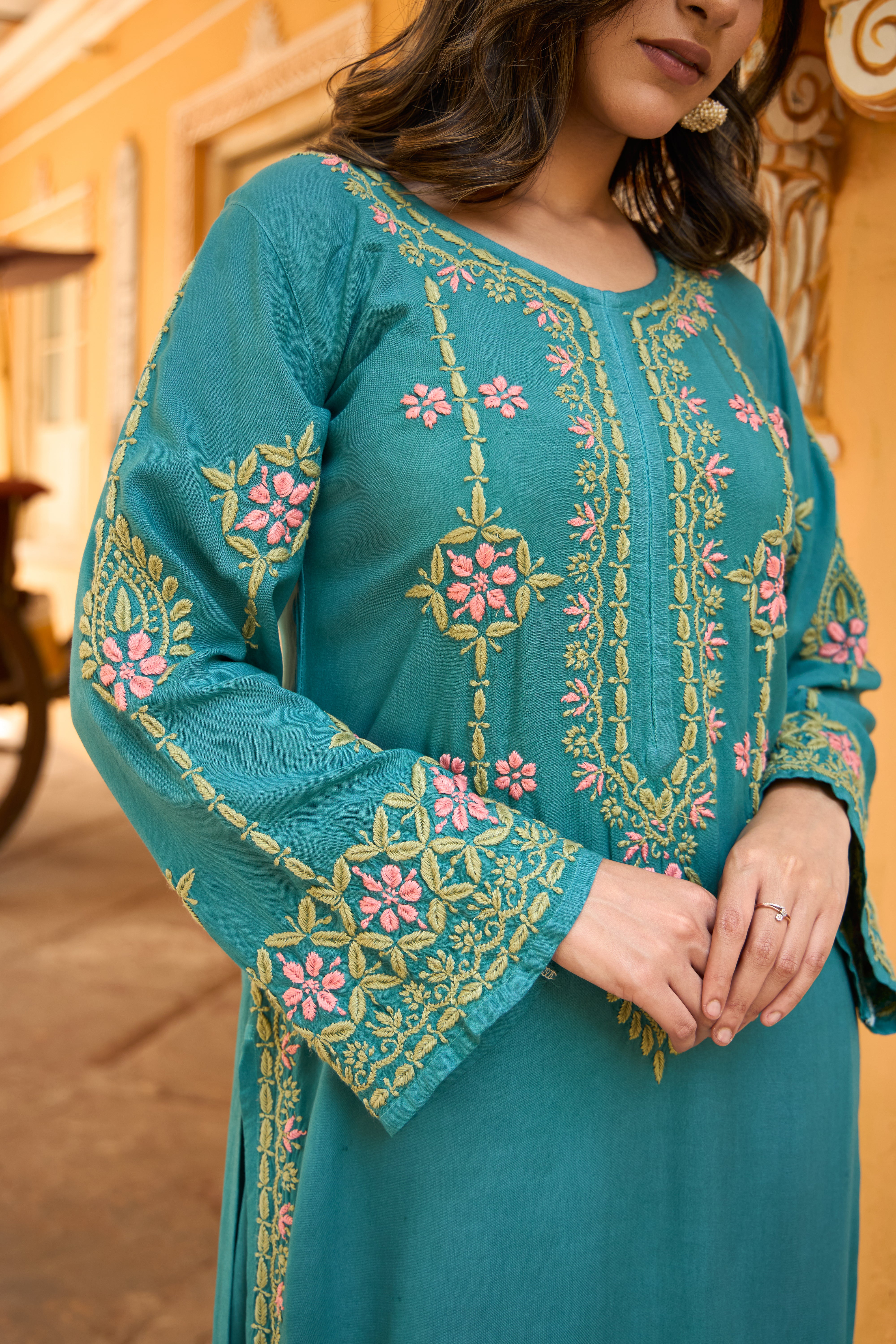 Teal embroidered dress with floral patterns worn by a person, against an ornate architectural background.