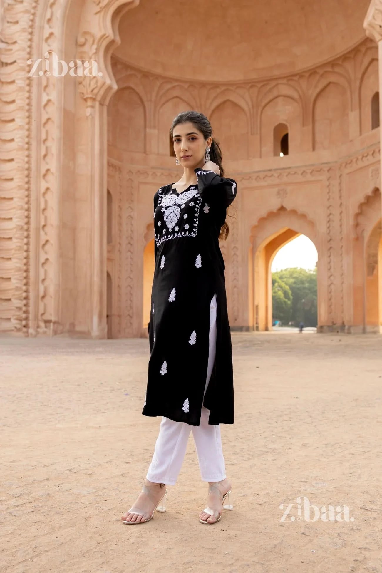 A woman posing in a beautiful black and white kurti with intricate embroidery, paired with white pants, in a grand heritage backdrop.