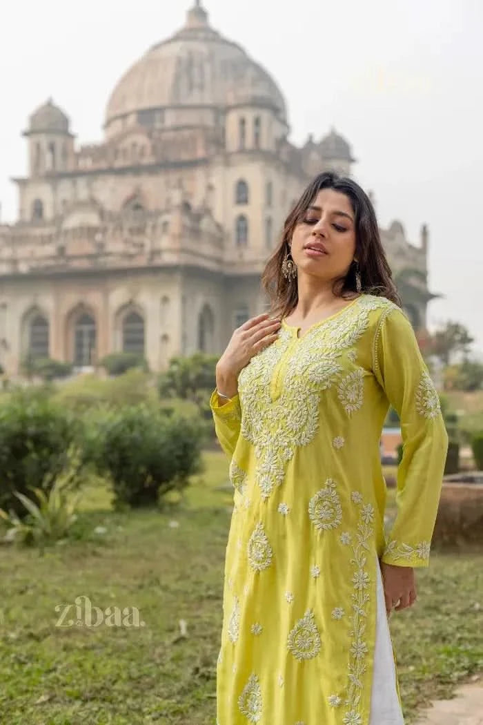A woman poses in a serene outdoor setting wearing a lime green Chikankari Kurti for women, adjusting her hair outside a historic monument.