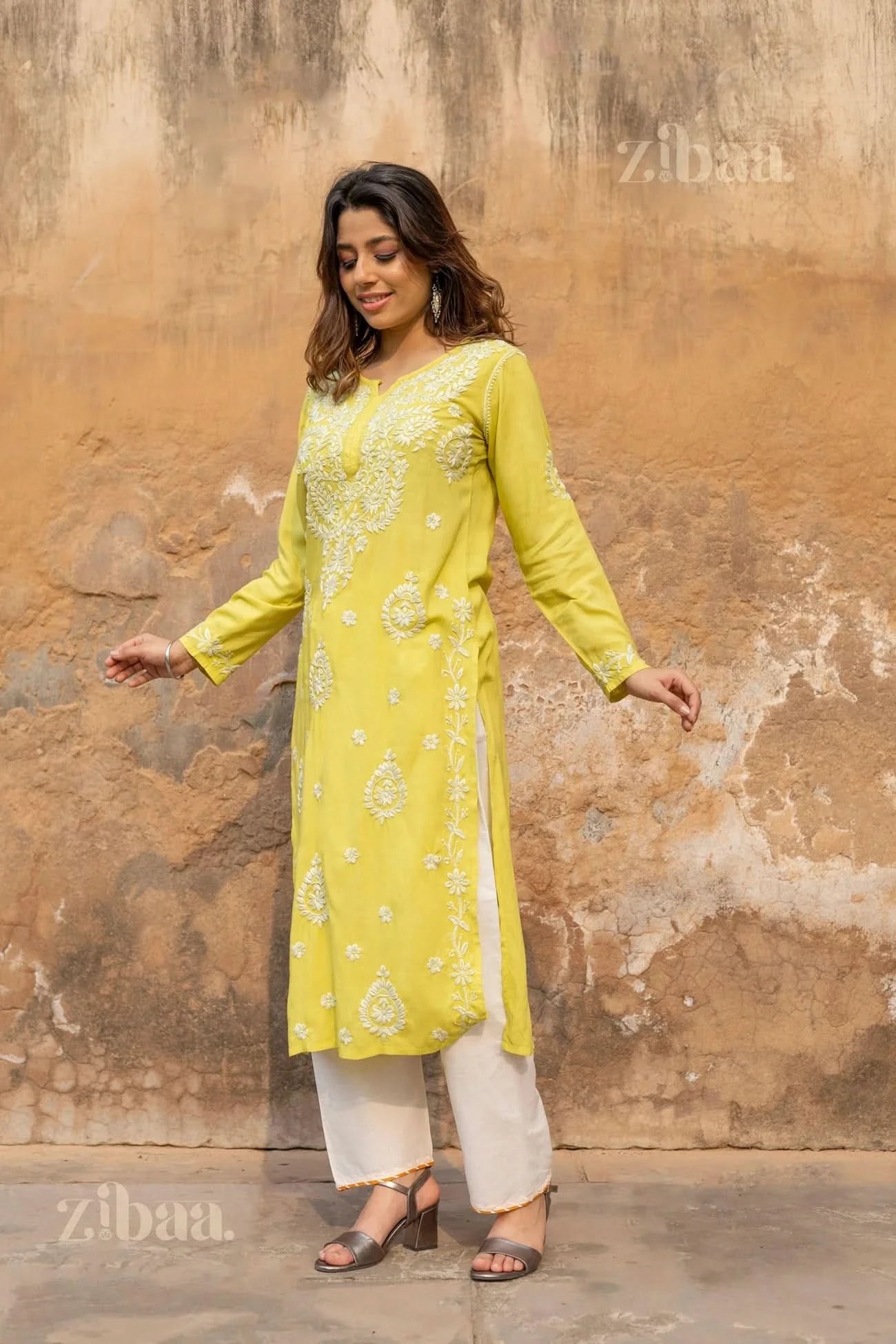 A woman stands outdoors in a lime green Chikankari long kurti, holding the sides of her kurti, paired with white pants and heels.