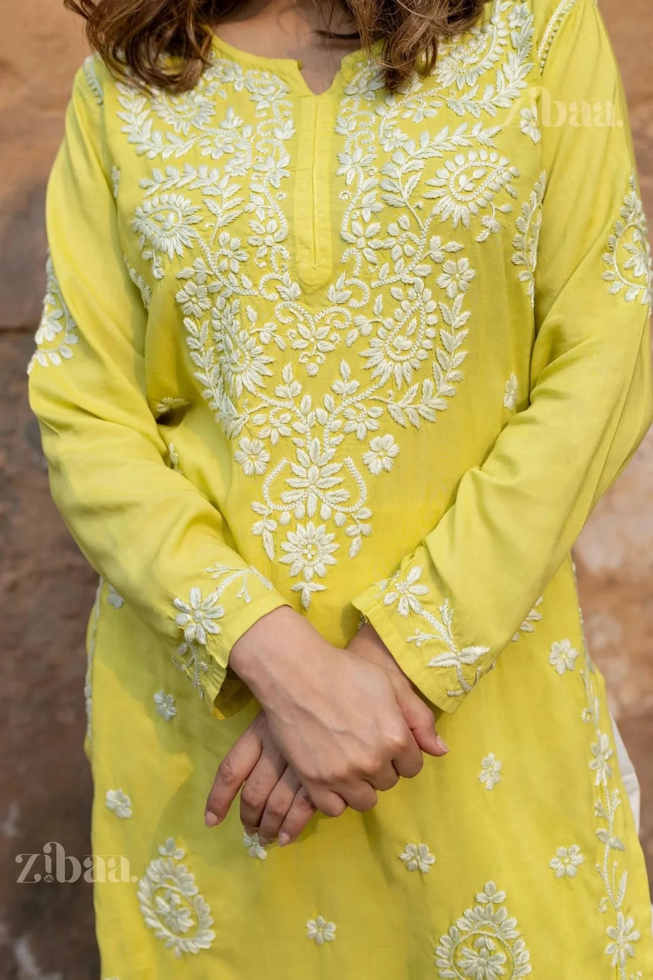 A close-up of a woman gently clasping her hands, wearing a lime green Chikankari Kurti with intricate white floral embroidery details.