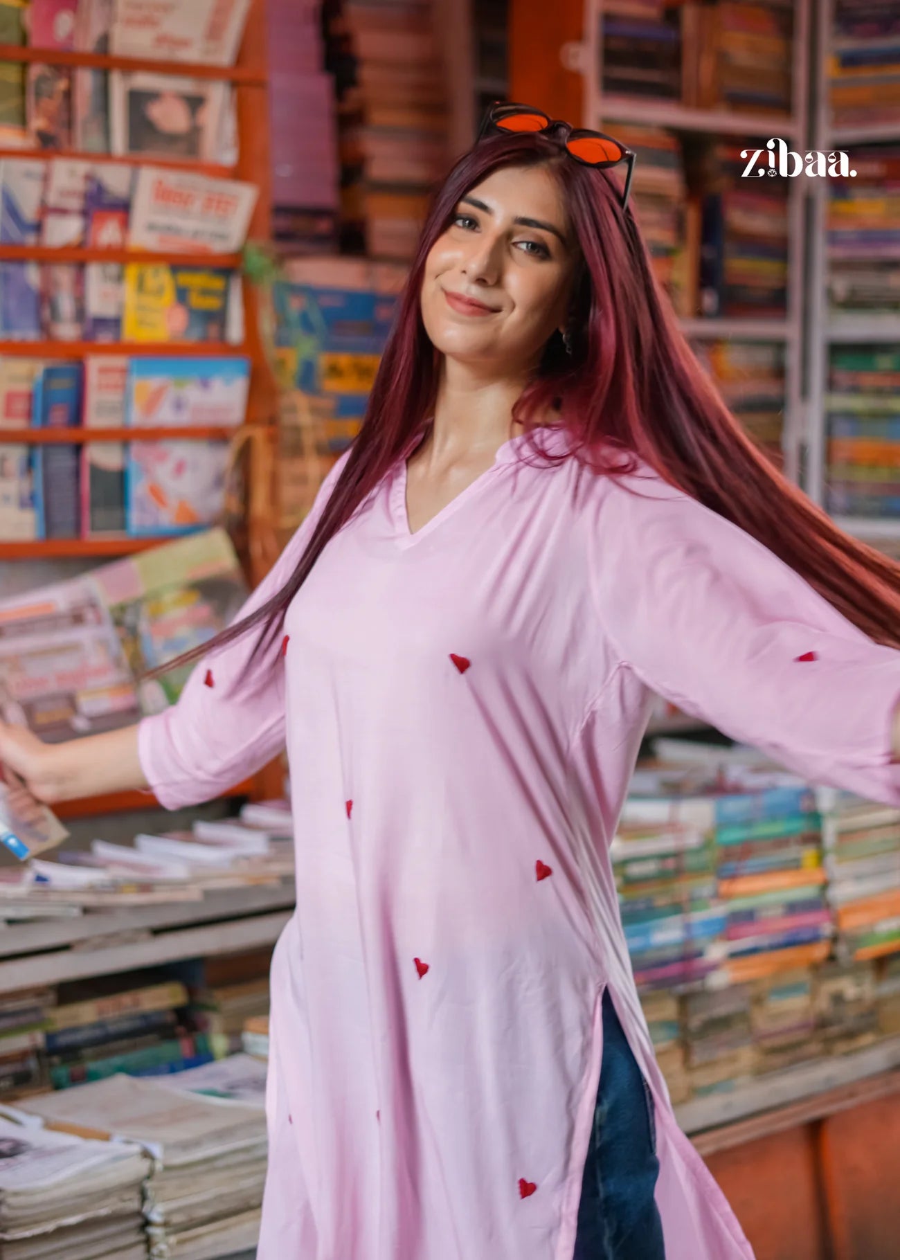 The model poses on a street with a book stall in the background, wearing a pink chikankari kurti adorned with heart motifs.