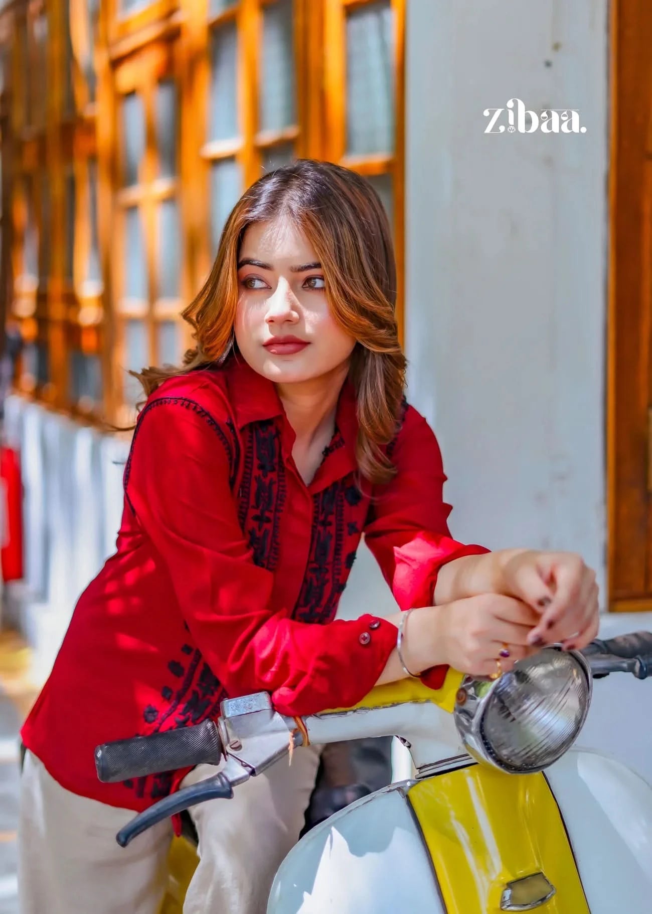 The model leans on the scooter, showcasing the fine black threadwork on her red chikankari shirt against an outdoor backdrop.