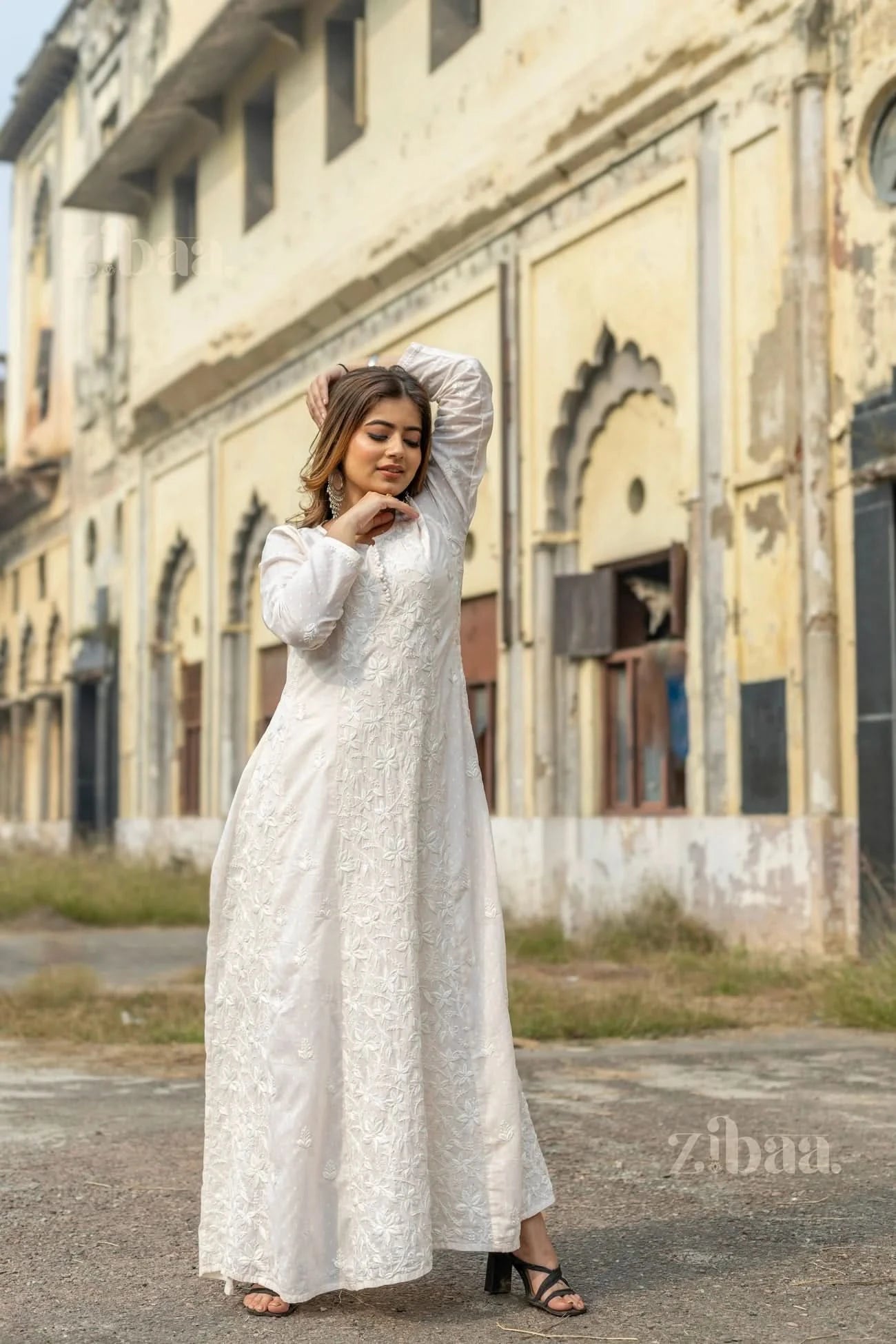 A woman wearing a white Chikankari kurti for women poses outdoors with one hand behind her head and the other resting on her neck.