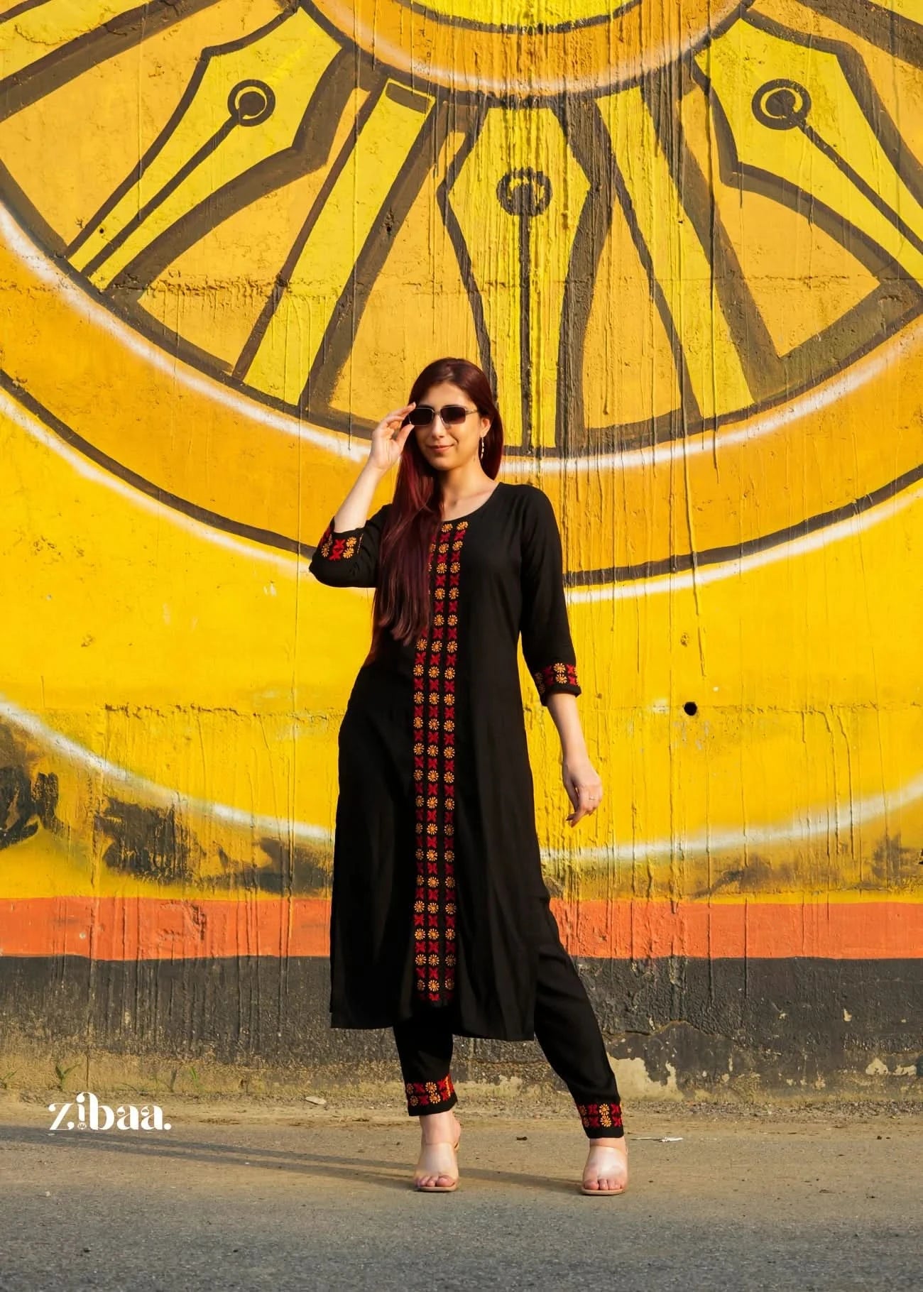 Woman in a black chikankari kurta set with red patterns standing in front of a yellow wall with circular designs.