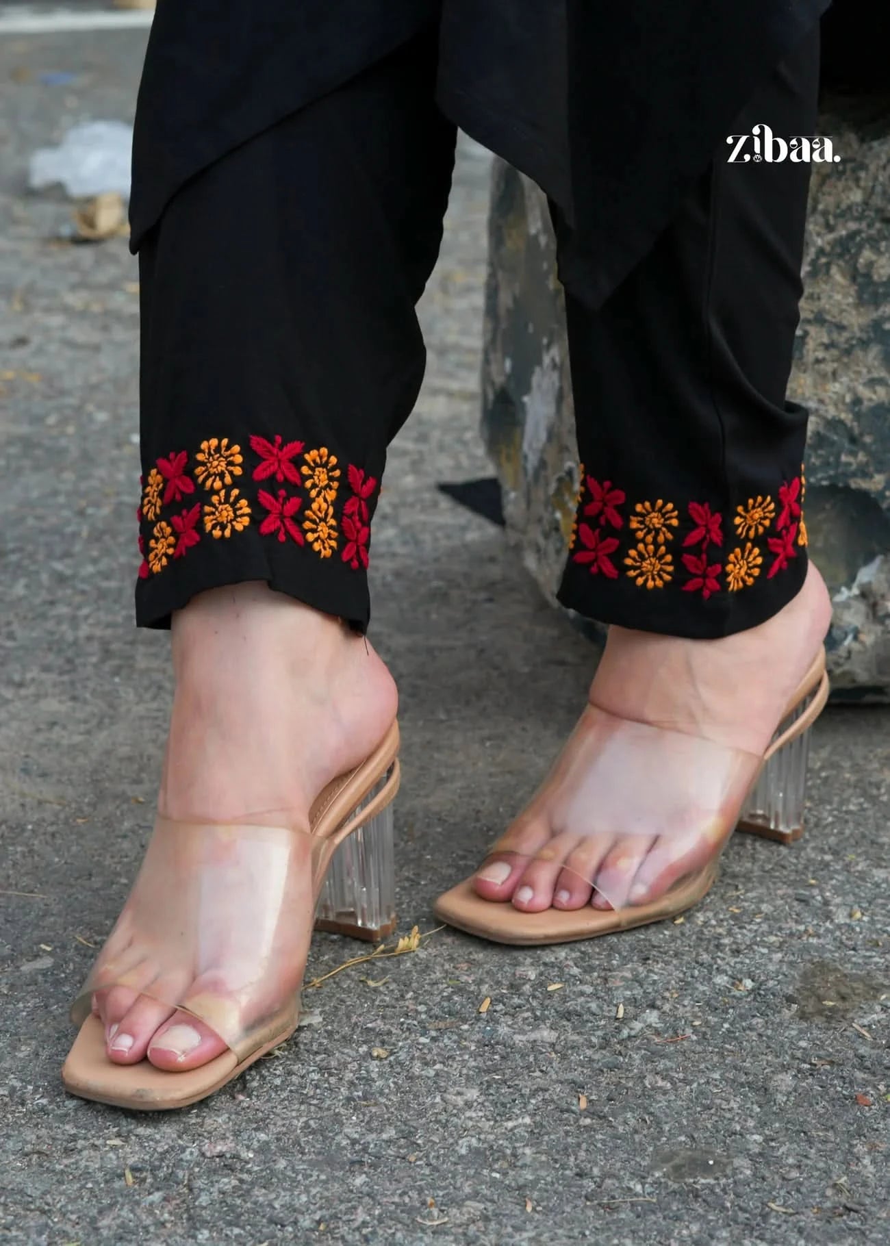 Black chikankari kurti set pants with colorful floral embroidery and beige sandals on a pavement.