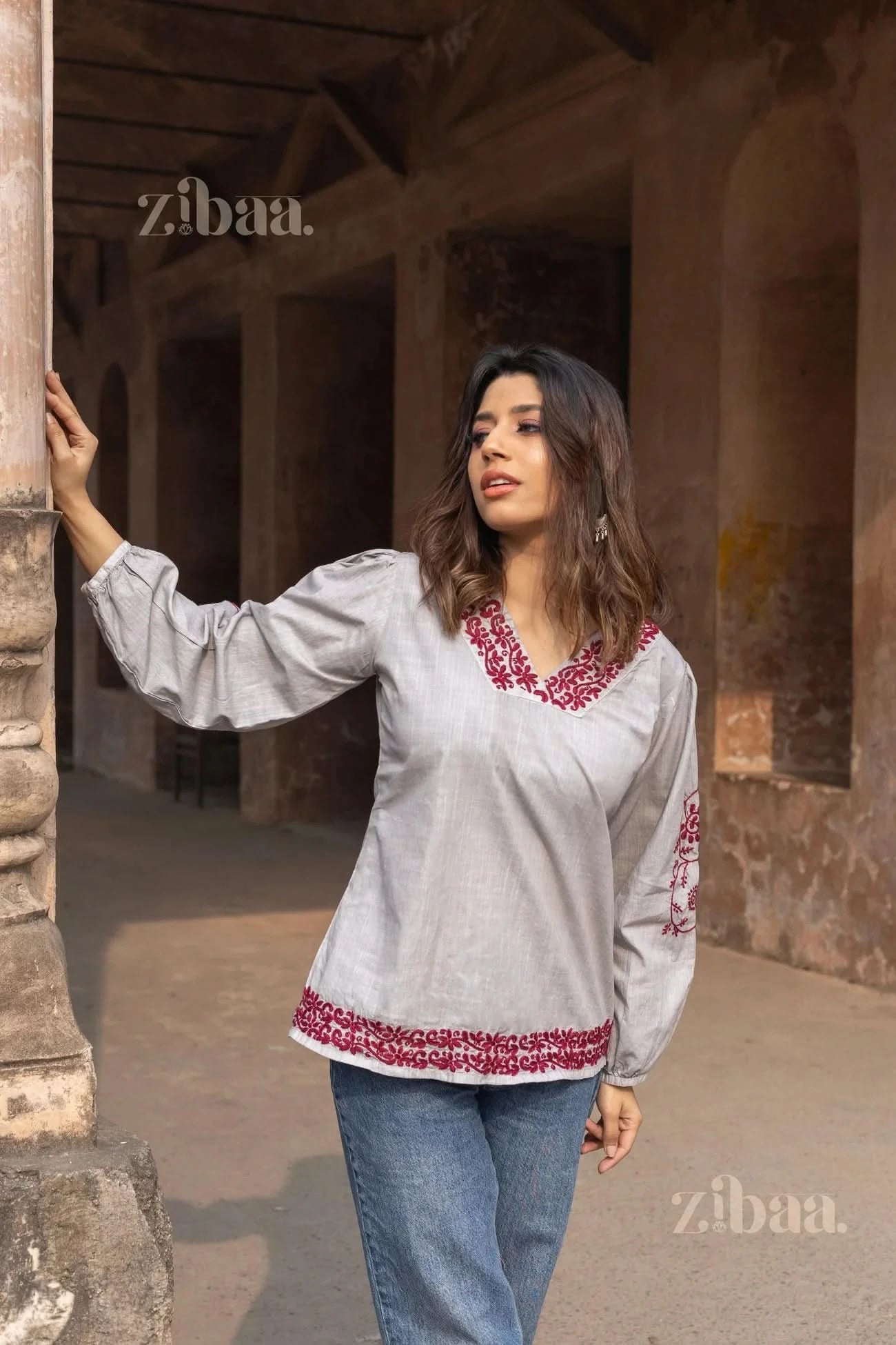 Woman standing outdoors, leaning against a pillar, wearing a grey Chikankari top with red embroidery on the neckline, sleeves, and hem.