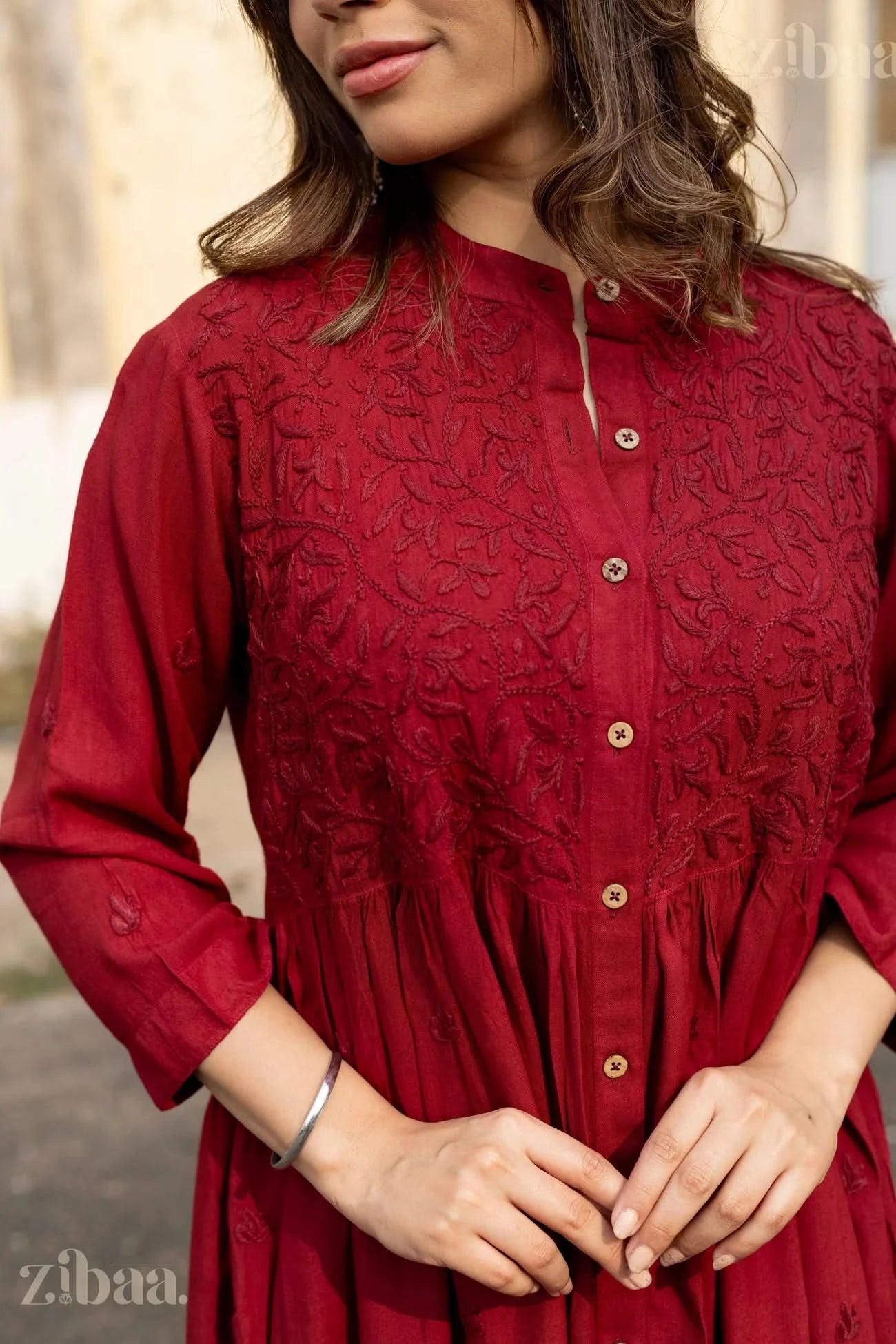 A woman gently holds the pleats of her maroon Chikankari top, showcasing tonal embroidery and button-down detailing in an elegant pose.