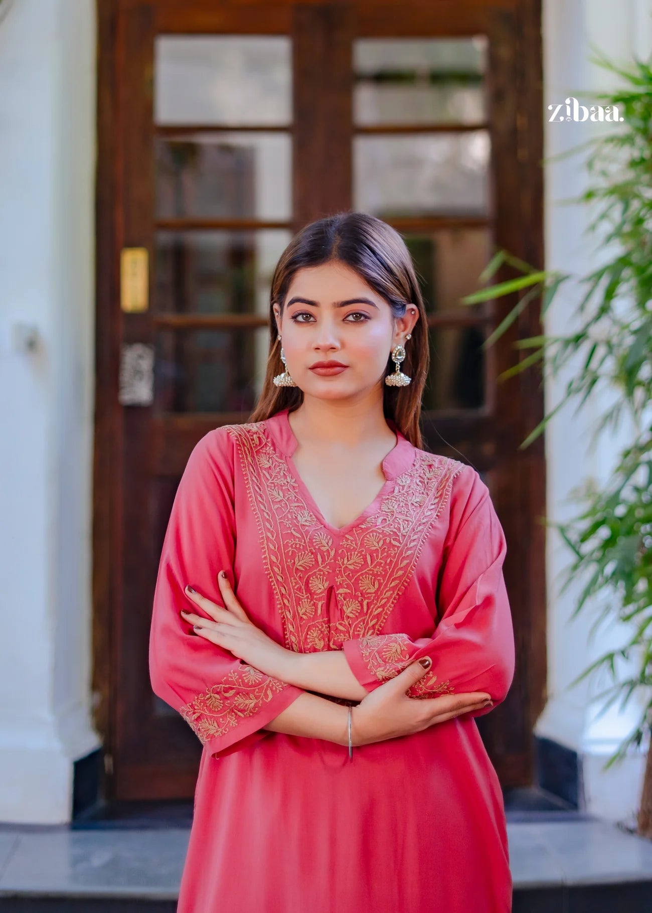 The model poses while standing, wearing a coral chikankari kurti with intricate stitching, framed by a rustic brown door behind her.