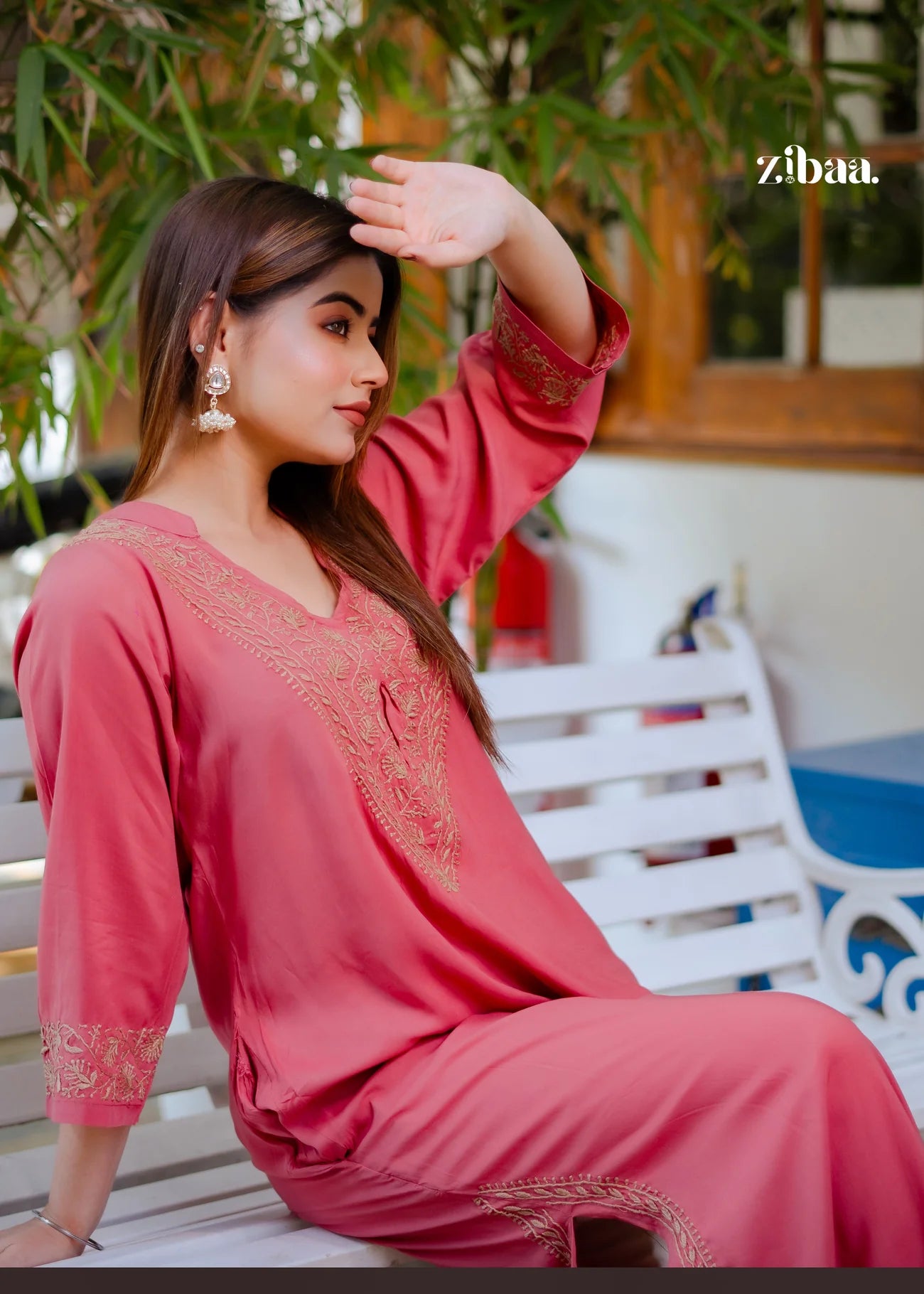 The model poses while sitting, wearing a coral chikankari kurti highlighted by its delicate golden embroidery.