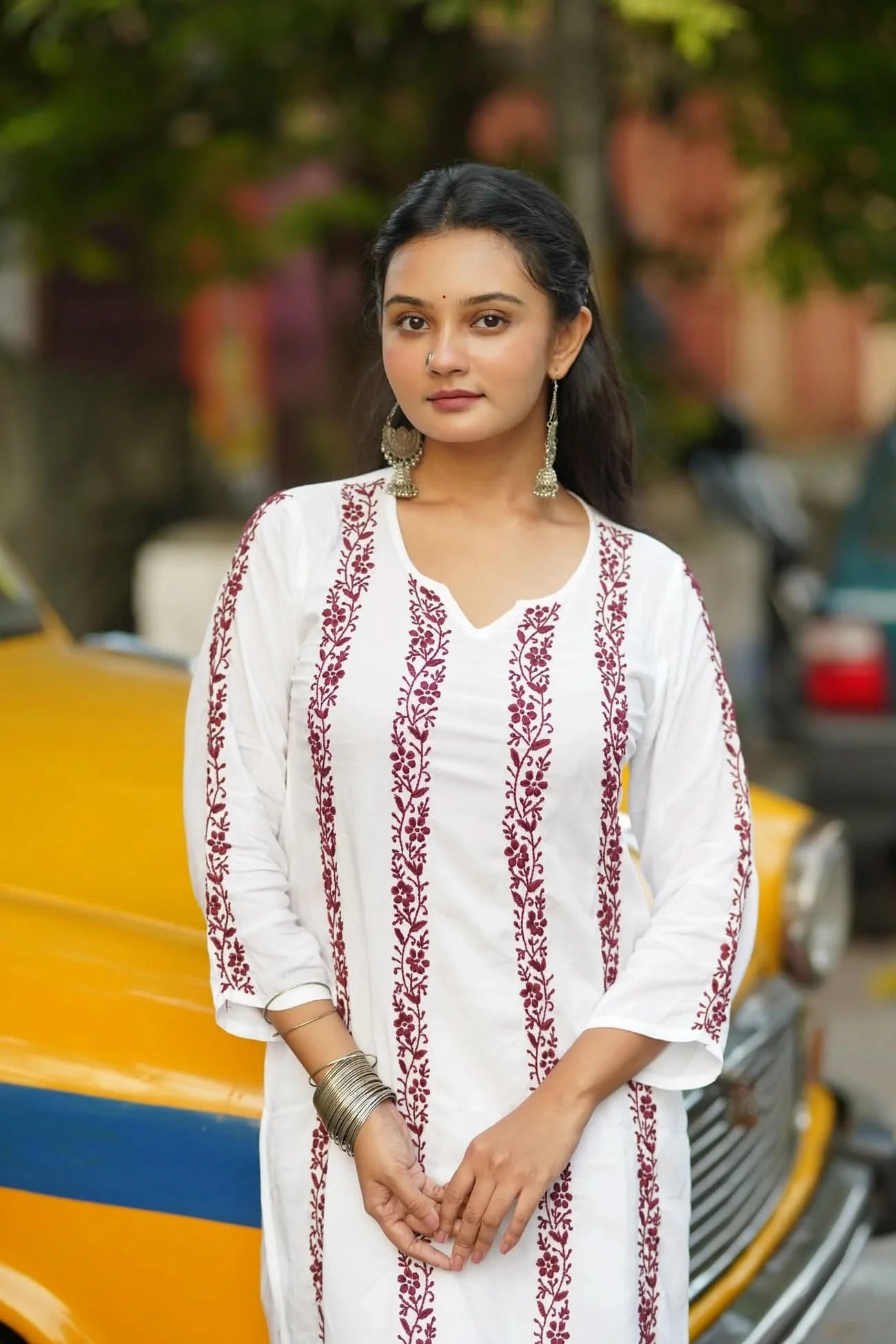 Woman in a white embroidered outfit standing in front of a yellow vehicle.