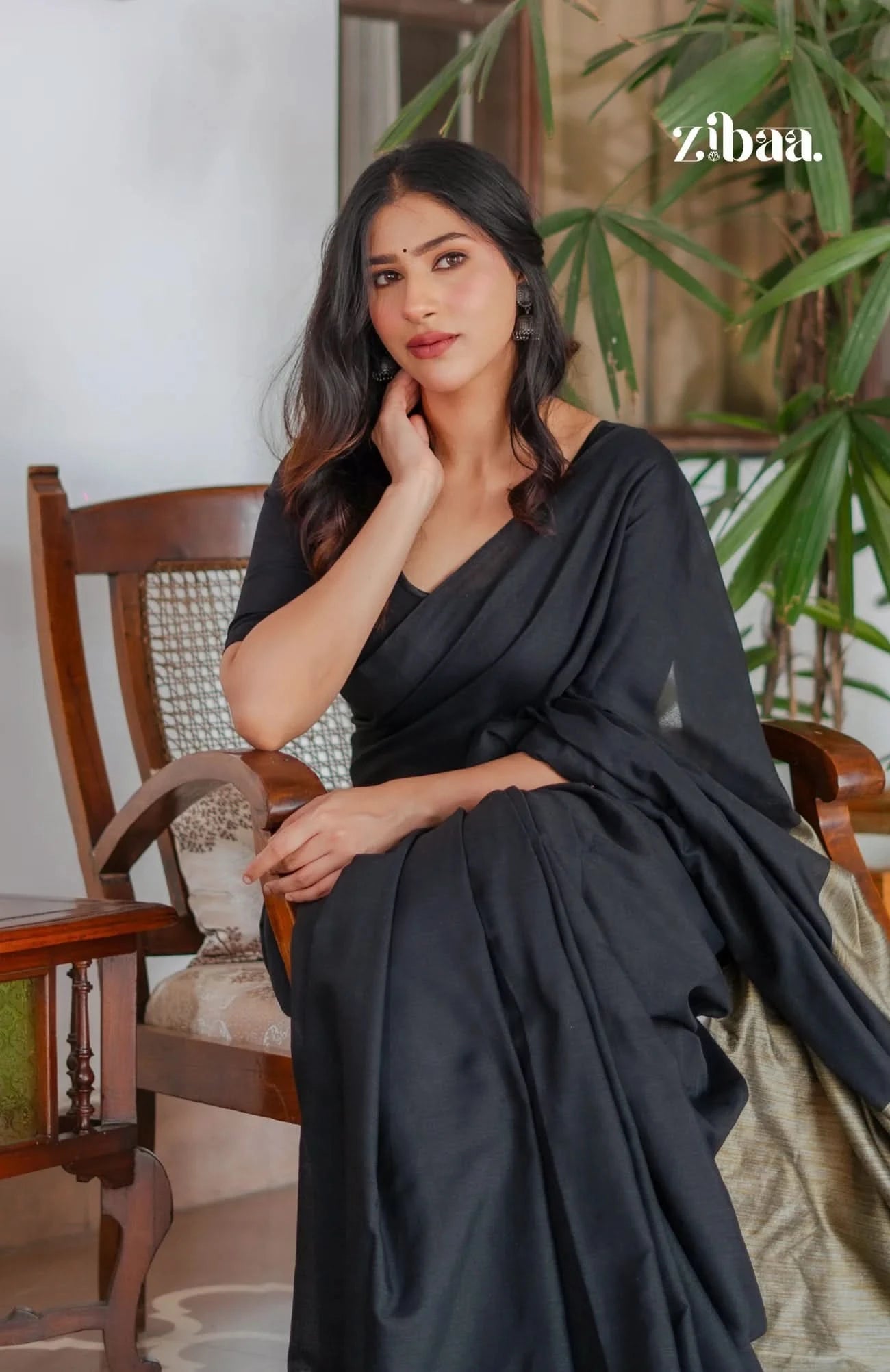 Woman in a black saree sitting on a wooden chair with a plant in the background, featuring the brand 'zibaa'.