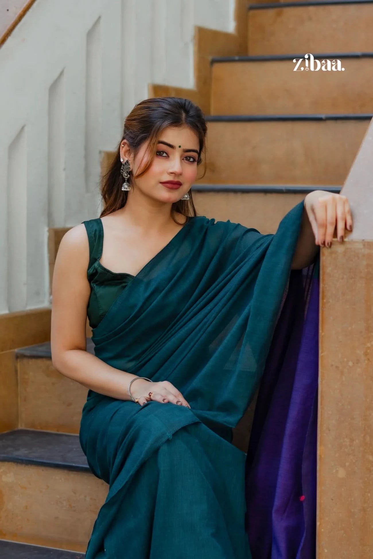 Woman in a teal saree sitting on stairs with 'zibaa' branding.
