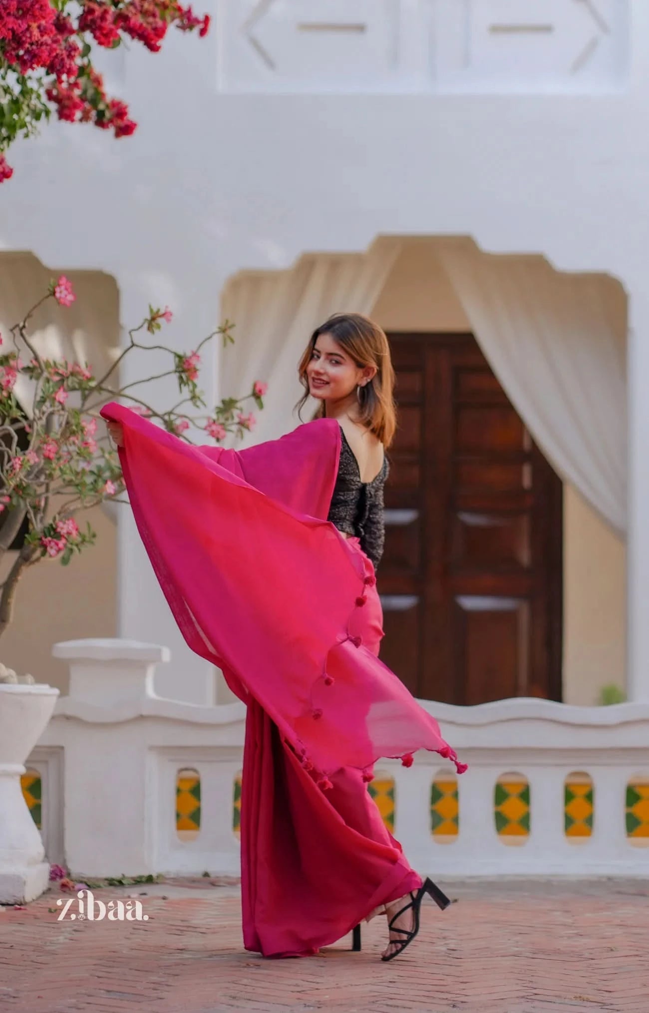 Woman in a pink saree standing in front of a decorative entrance with flowers.