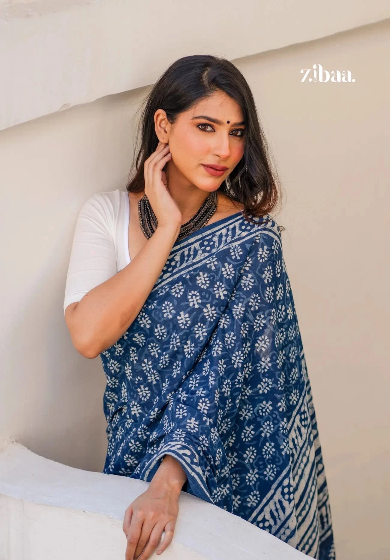 Woman wearing a blue and white patterned saree sitting on a beige couch, with 'zibaa' branding visible.